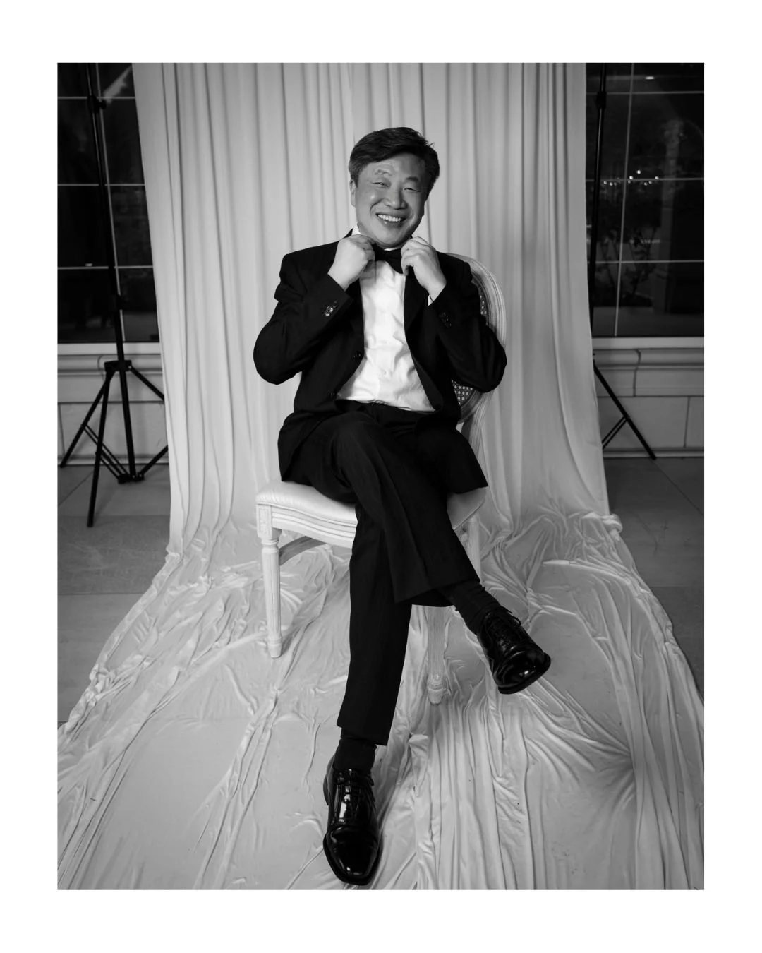 Smiling man in a tuxedo adjusting his bow tie, seated in front of a draped backdrop