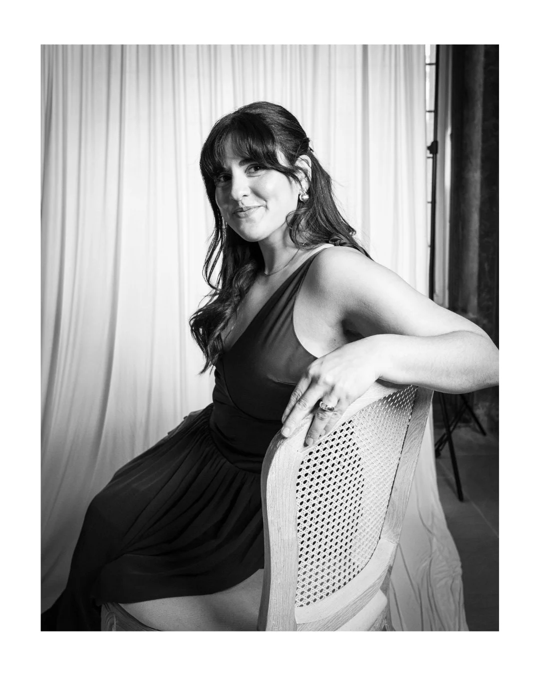 Black and white portrait of a smiling wedding guest in a dark dress, seated on a cane-back chair in front of a white draped backdrop