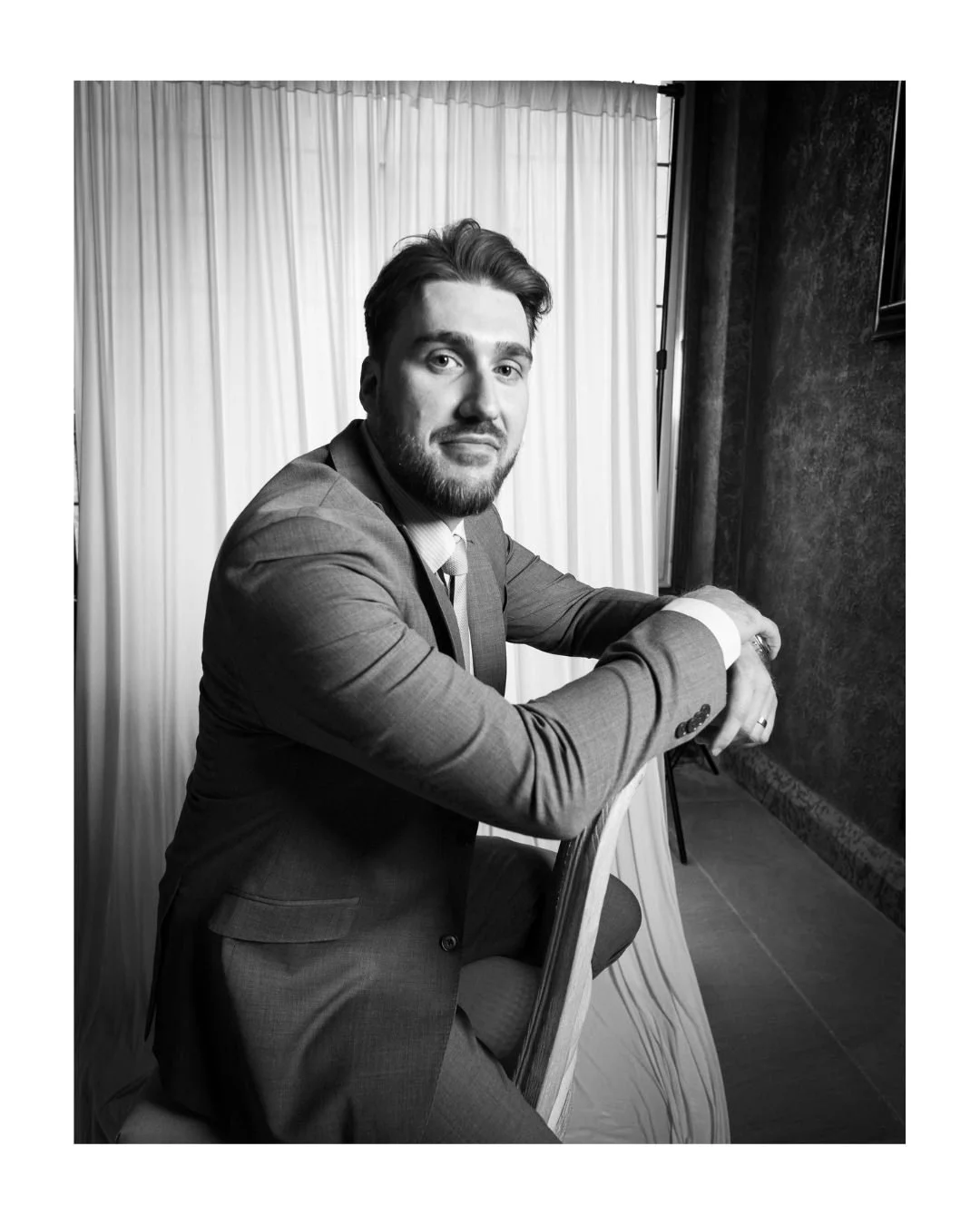 Black and white portrait of a man in a suit seated in front of a draped backdrop and textured wall