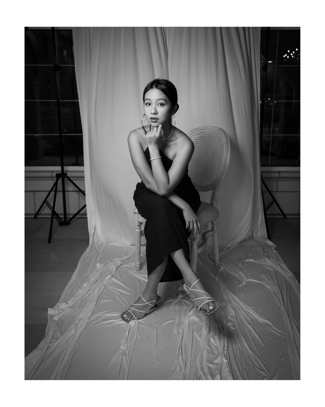 Black and white portrait of a young woman in hoop earrings, seated on a cane-back chair with her chin on her hand