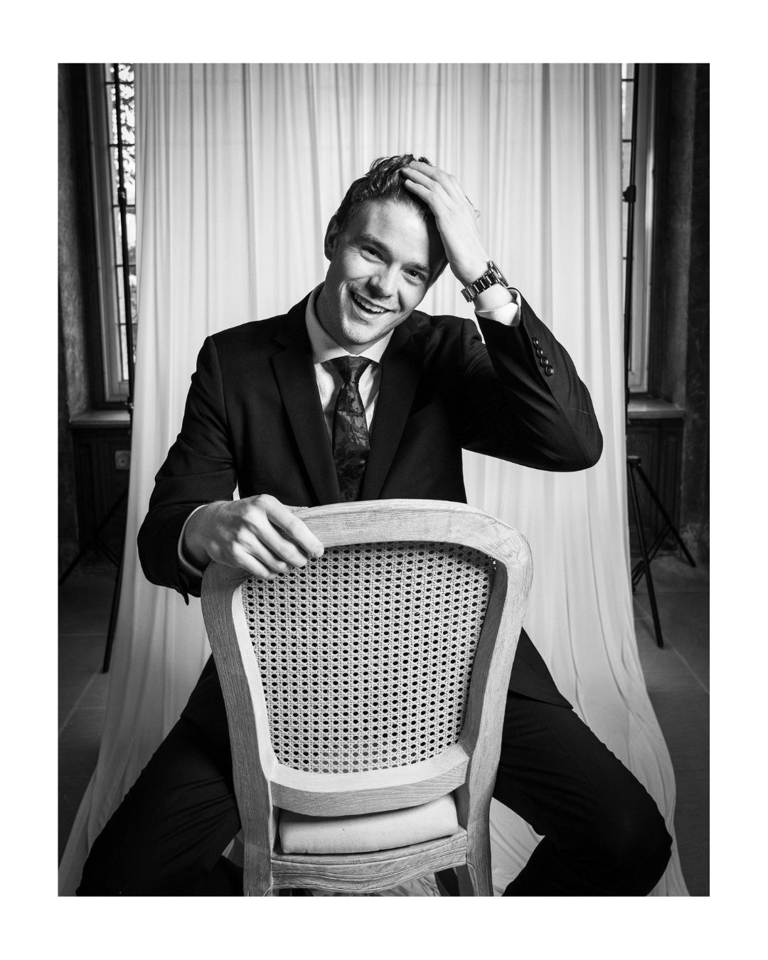 Smiling man in a suit and tie leaning on a chair back, hand on his head, black and white portrait