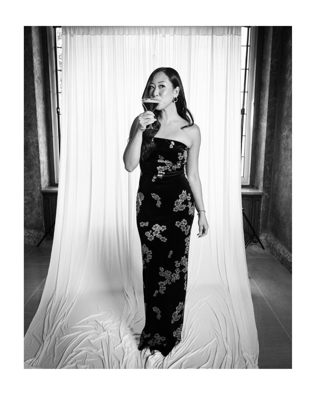 Wedding guest in a floral strapless gown holding a cocktail in front of a light draped backdrop