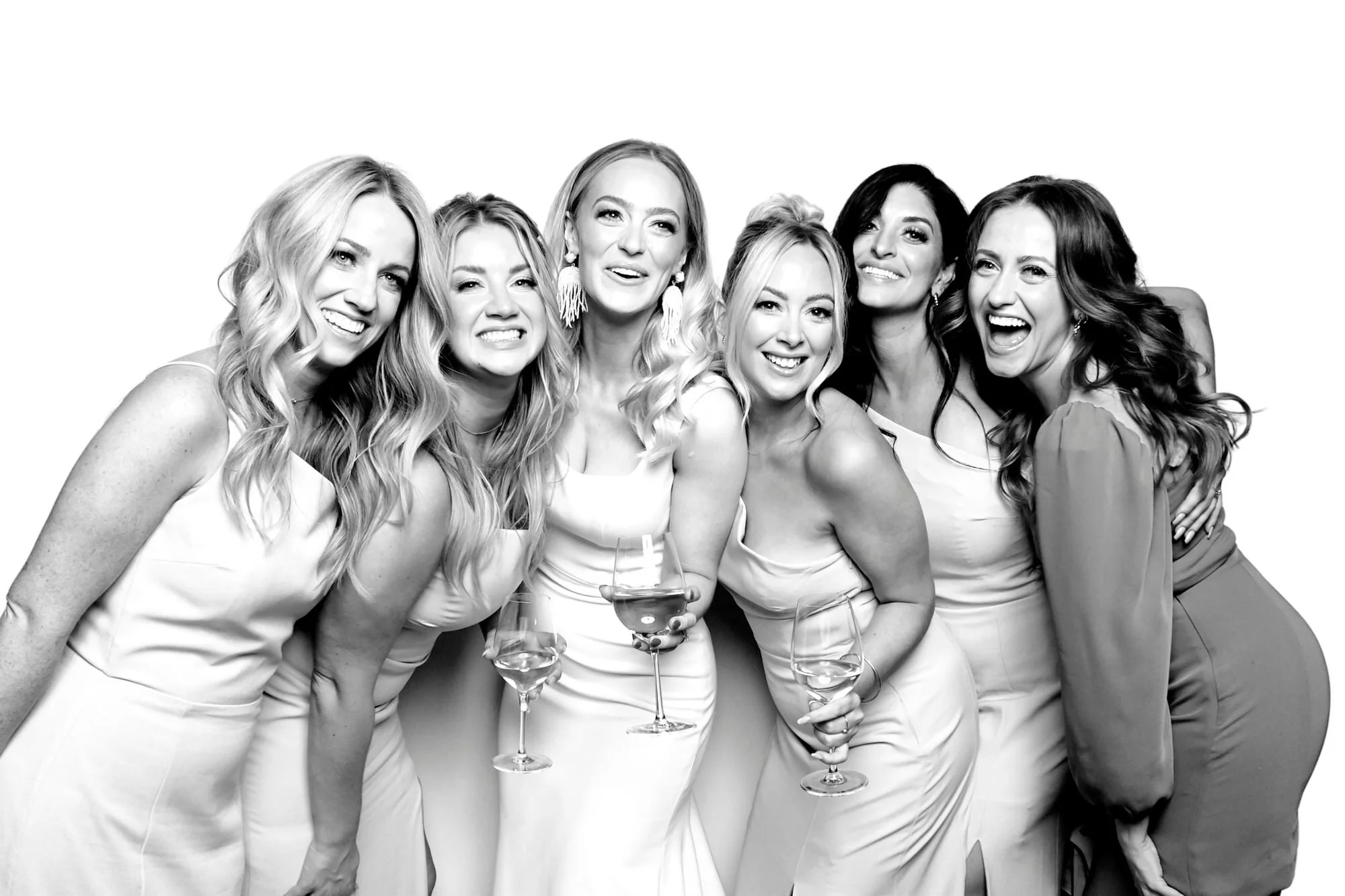 Seven laughing women in light dresses holding wine glasses, black and white group photo on a white backdrop