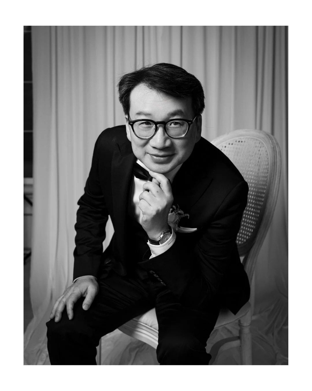 Black and white portrait of a man in a tuxedo and glasses seated on a woven chair