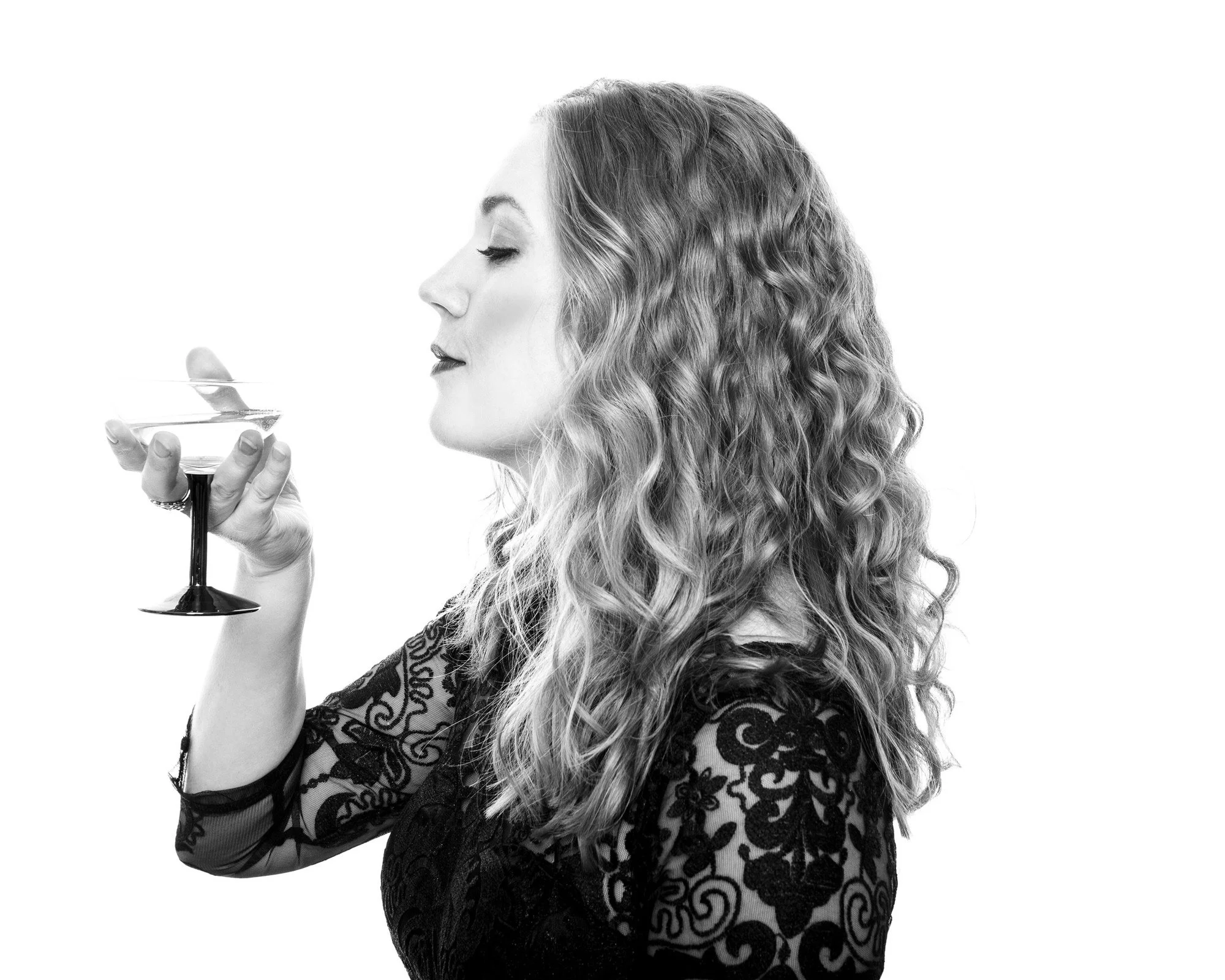 Profile portrait of a woman in a black lace dress holding a coupe glass, eyes closed, on a white backdrop