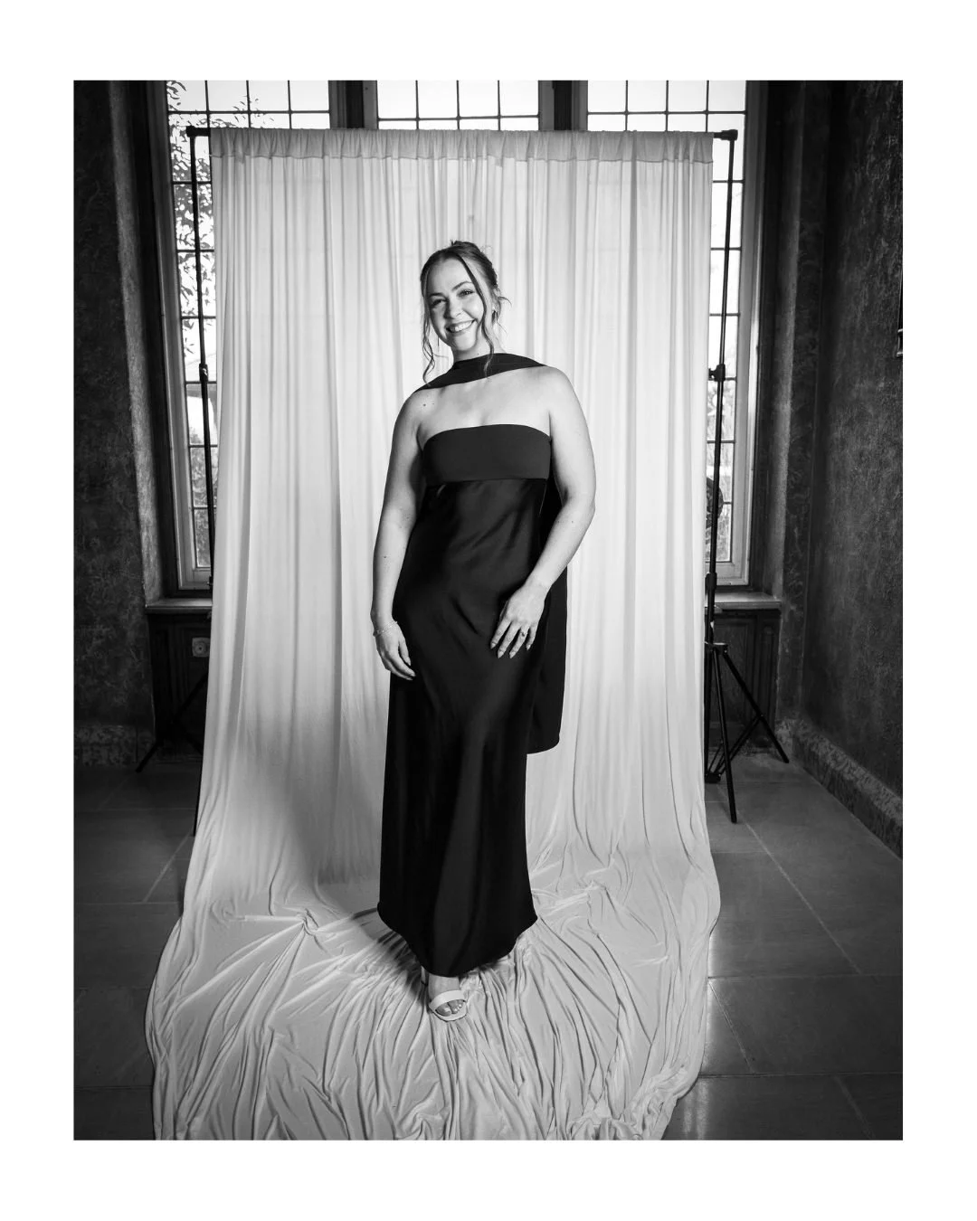 Smiling wedding guest in a black evening gown in front of a white draped backdrop