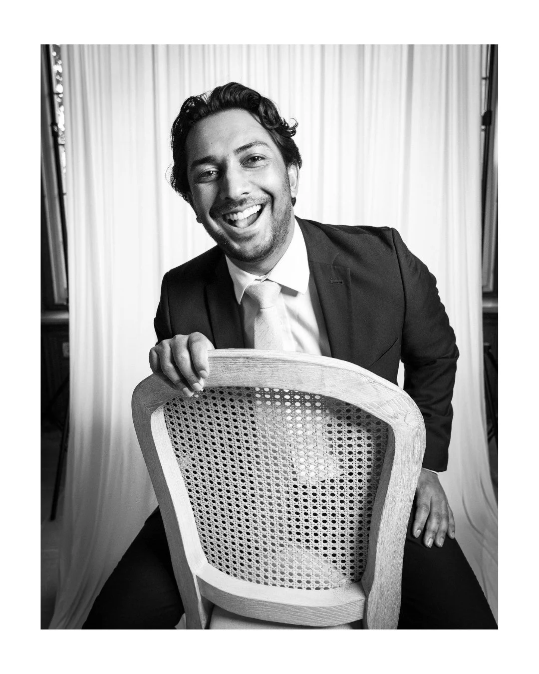 Black and white portrait of a laughing man in a suit holding a cane-back wooden chair