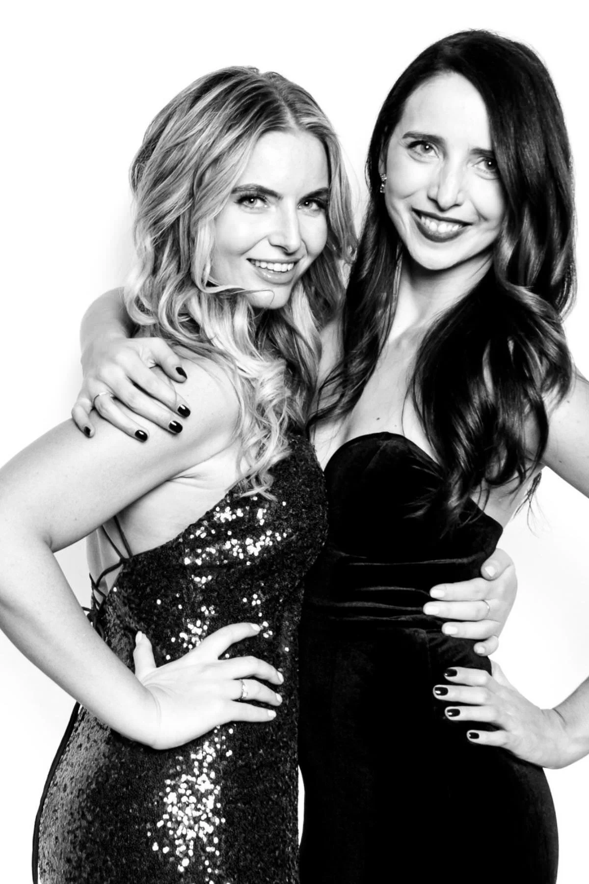Two smiling women, one in a sequinned dress and one in black velvet, with arms around each other