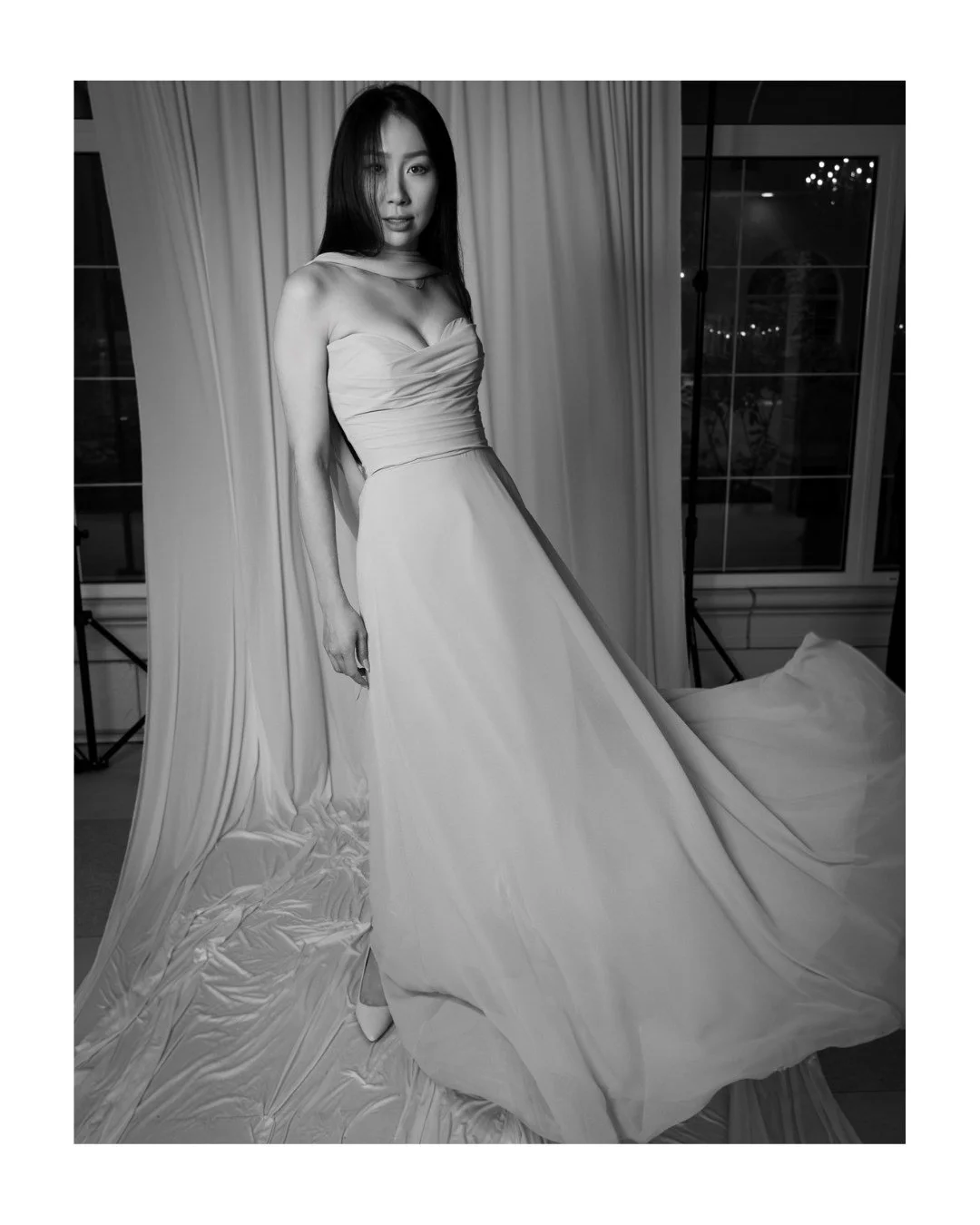 Black and white portrait of a woman in a strapless flowing gown in front of a draped backdrop, with a night window behind