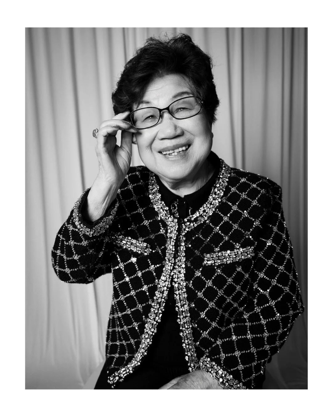 Black and white portrait of a smiling older woman in a sequined jacket adjusting her glasses in front of a draped backdrop