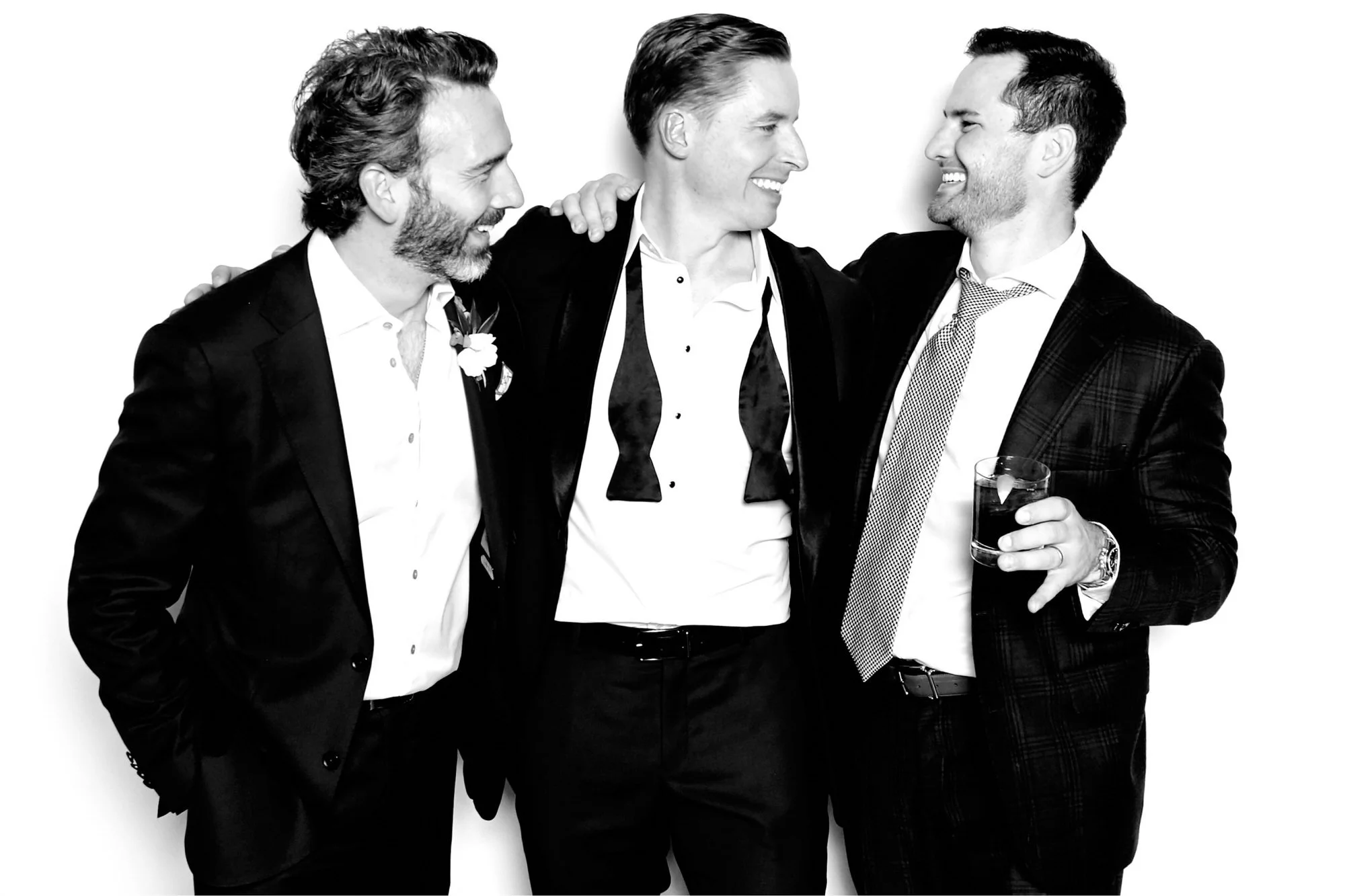 Three laughing men in suits, one with an untied bow tie and one with a boutonniere, on a white backdrop