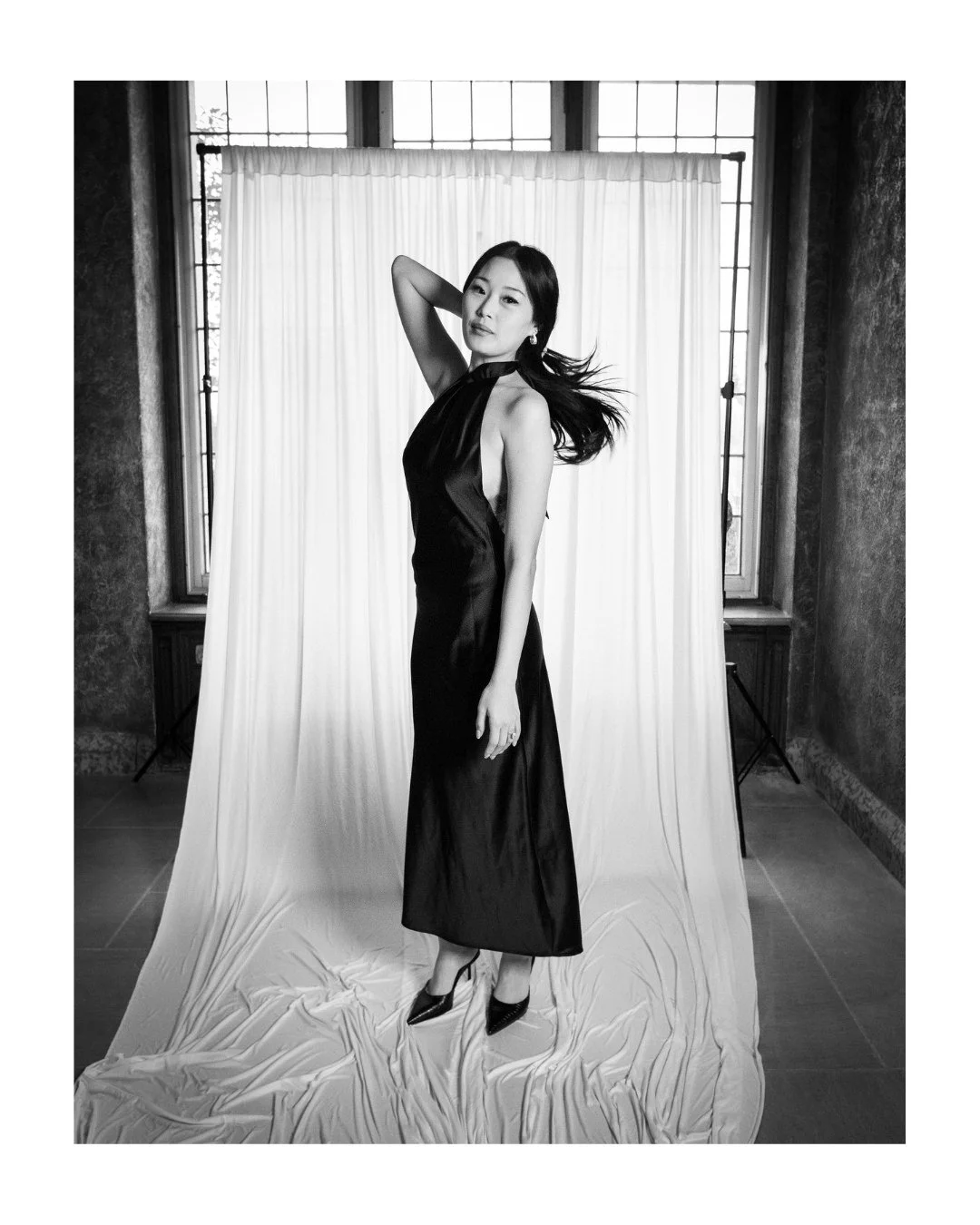 Wedding guest in a long gown posing with one hand behind her head in front of a white draped backdrop