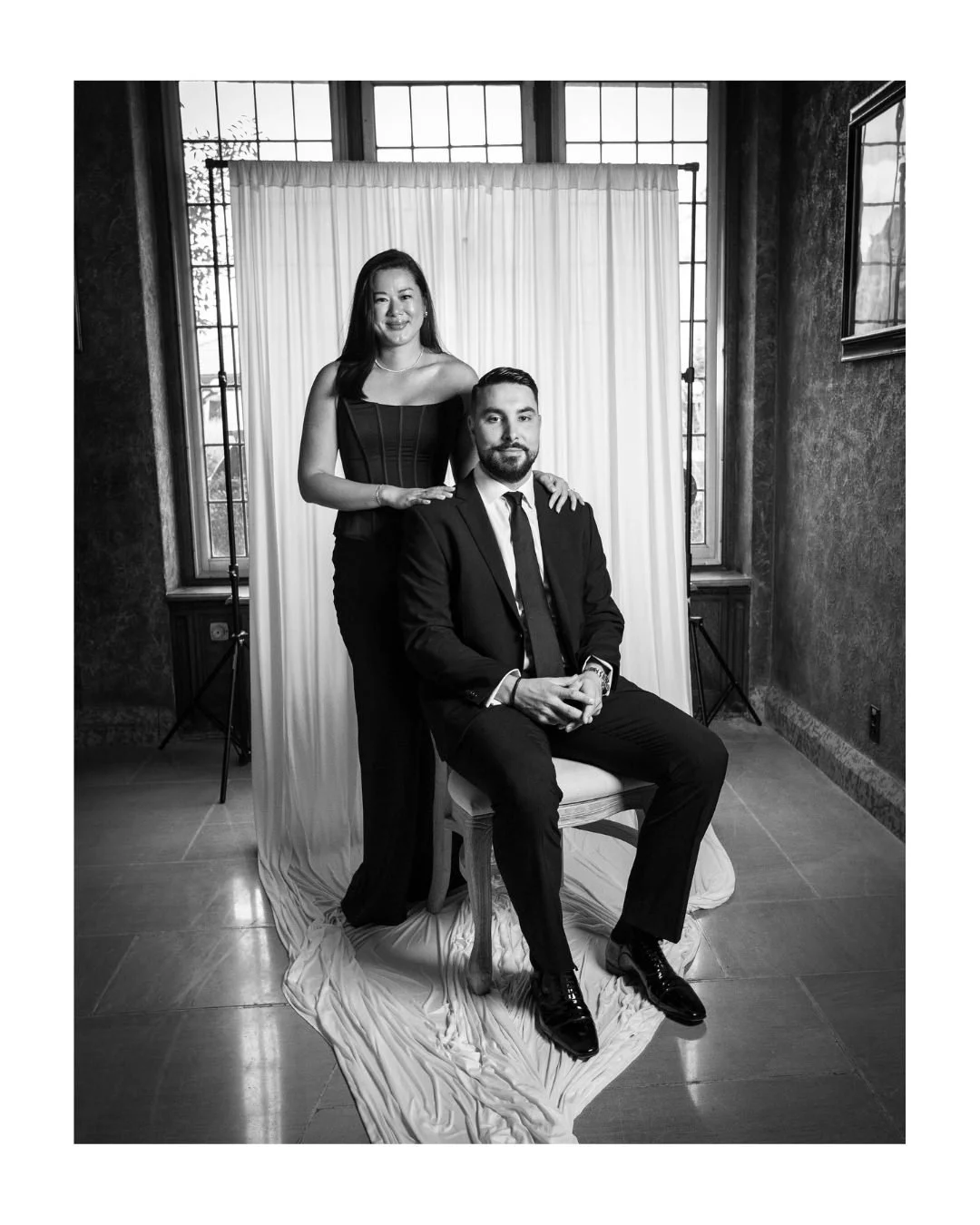 Black and white portrait of a woman standing behind a seated man in a suit in front of a draped backdrop