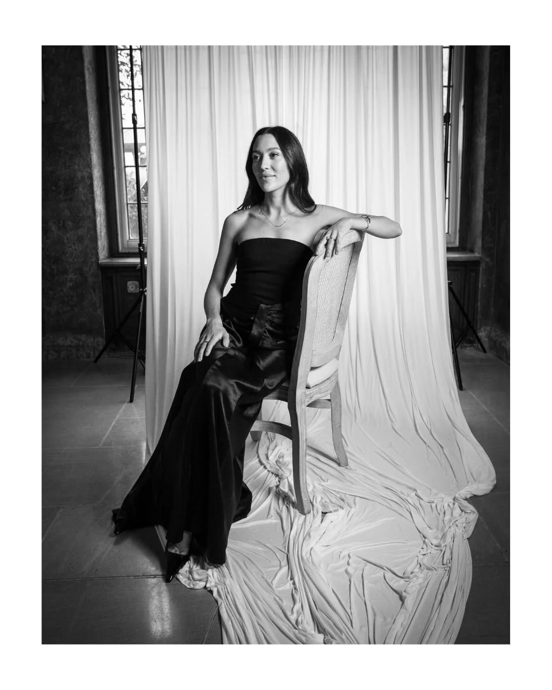 Black and white portrait of a woman in a strapless dress seated on a wooden chair in front of a draped backdrop