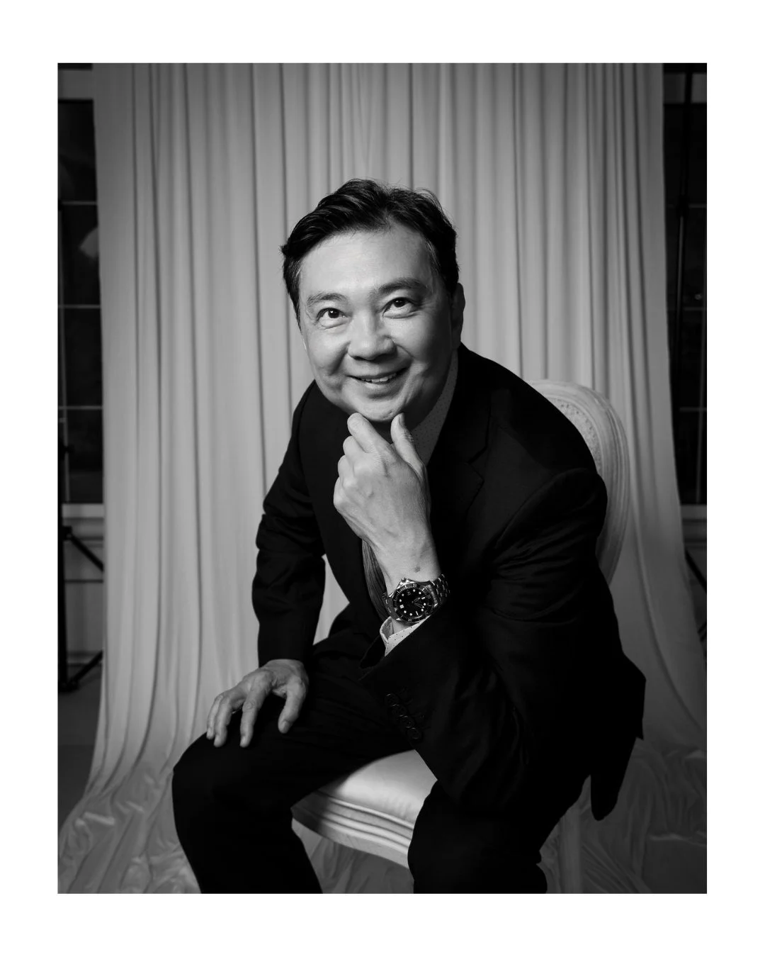 Black and white portrait of a smiling man in a suit seated with his hand resting on his chin