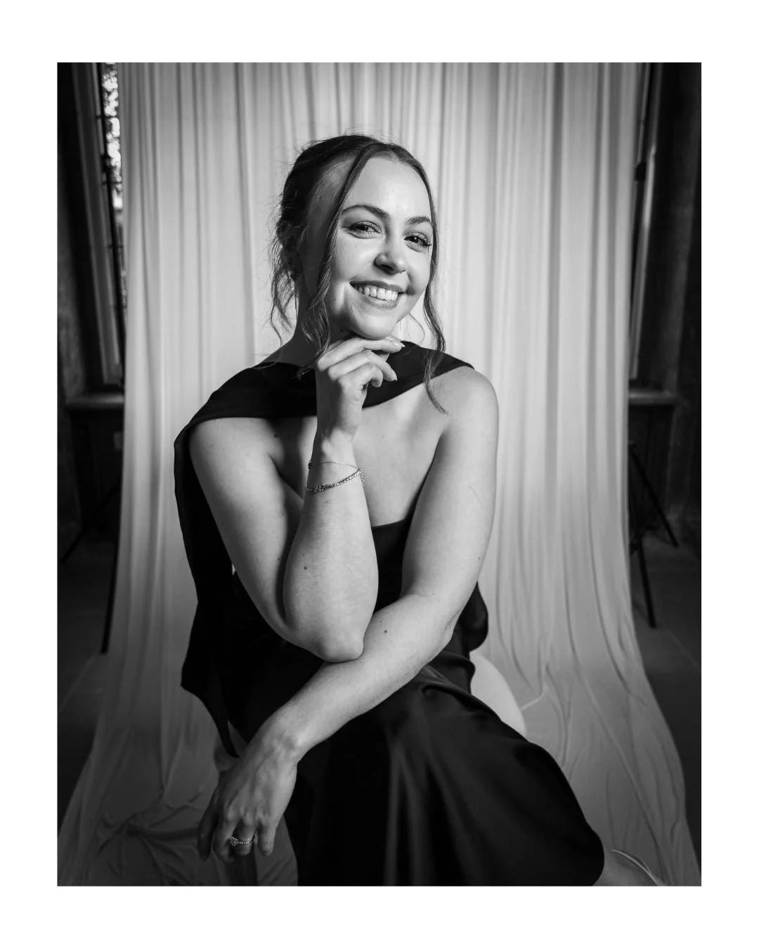 Black and white portrait of a smiling woman resting her chin on her hand in front of a draped backdrop