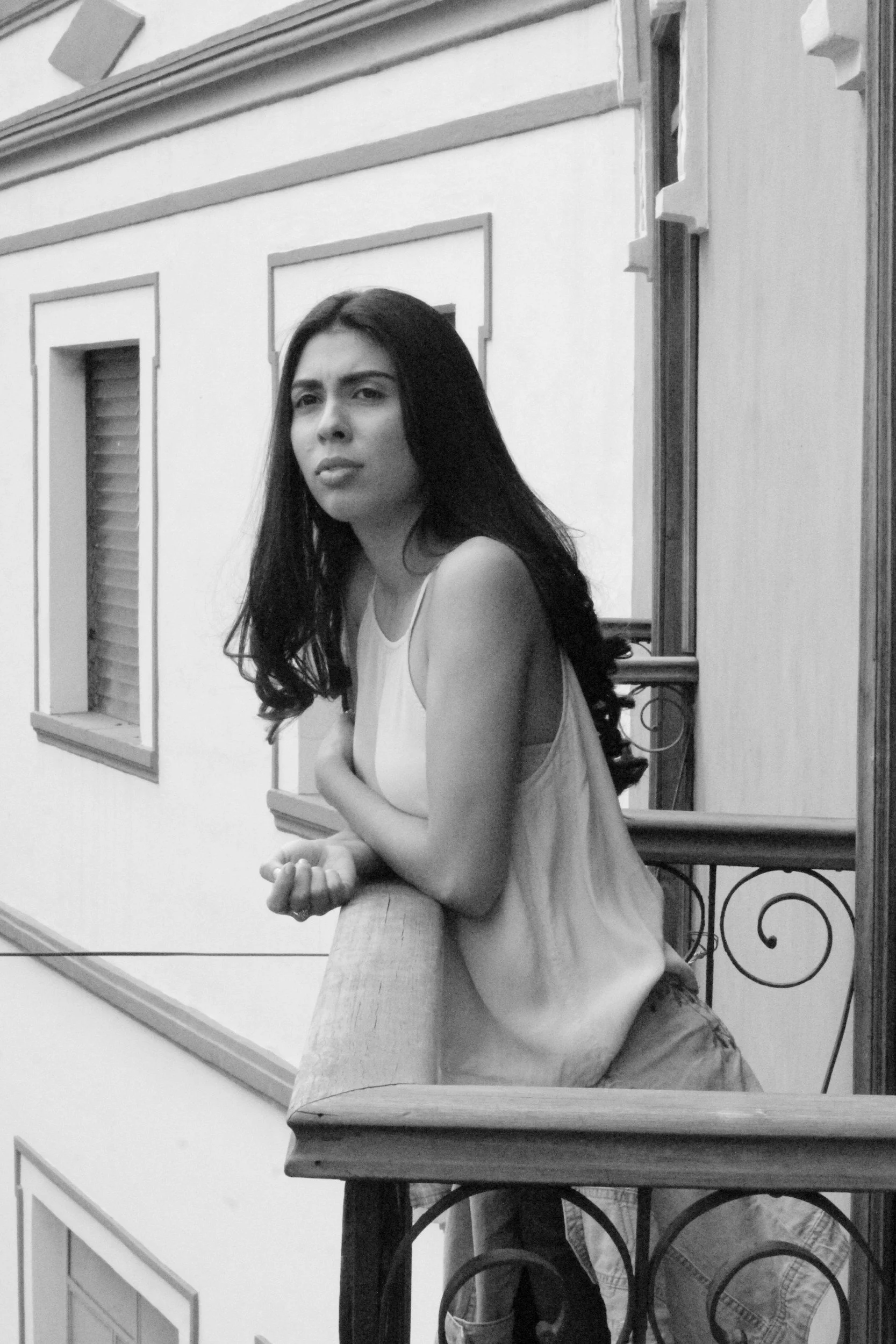 Black and white photo of a young woman with long dark hair, sitting at a table, close-up, with a serious pensive facial expression, in front of a decorative wall with framed windows longing for peace.