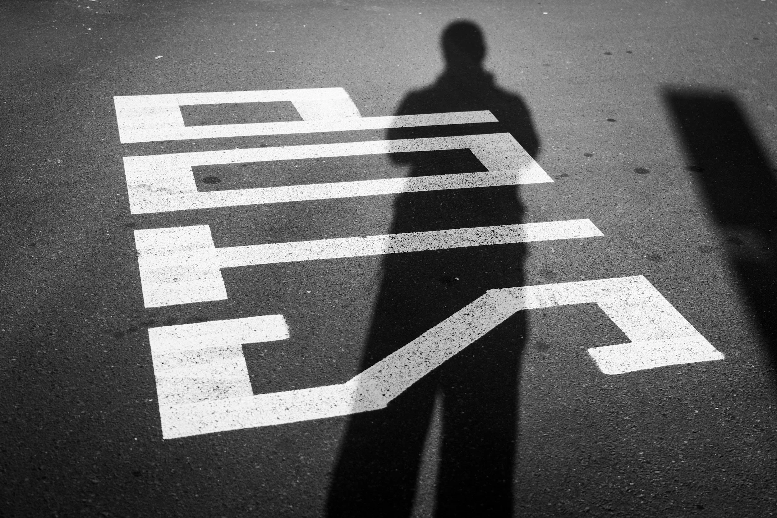 Shadow of a person on a crosswalk painted on the road, with the person's head and shoulders visible in the shadow exploring boundaries.
