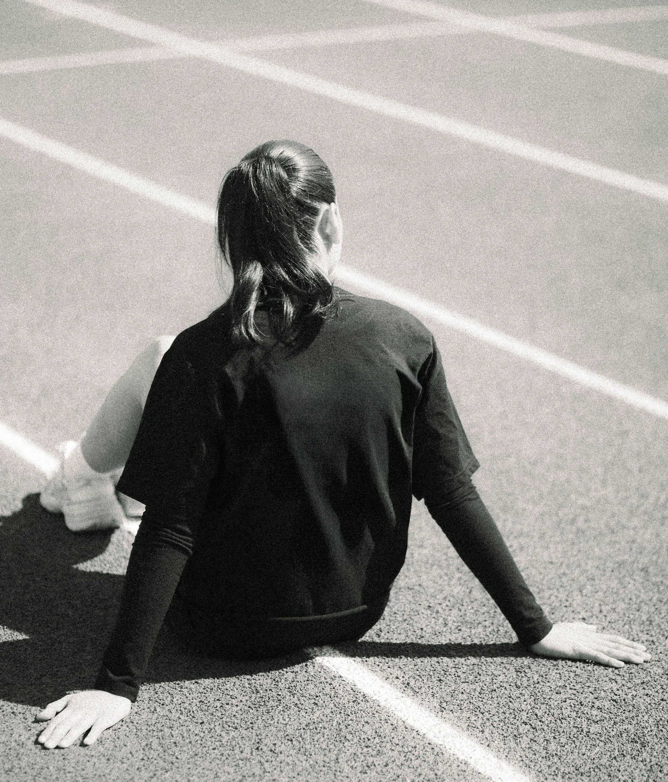 An athlete in black athletic wear is positioned on one side of a racetrack at night, with her hair in a braid, sitting on the ground with her hand touching the pavement. The racetrack has white lane markings, and the image is in black and white.