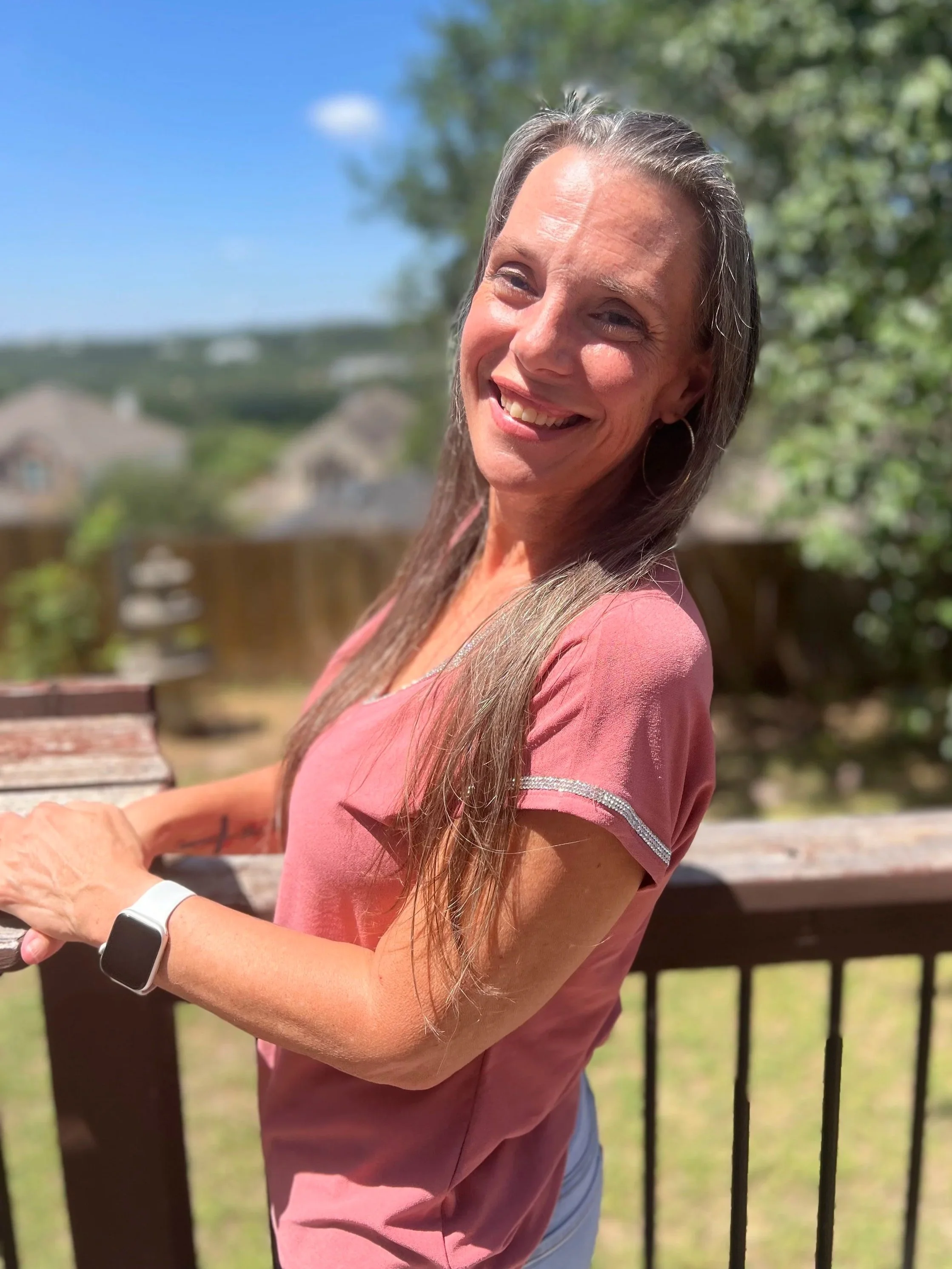 A smiling woman with gray hair and hoop earrings outdoors, wearing a pink shirt and a smartwatch on her left wrist, standing by a wooden fence with a background of trees and houses under a blue sky.