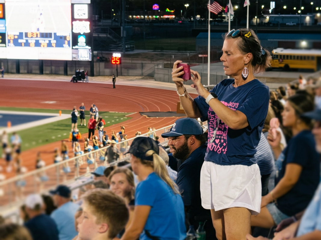Angela Allen, LPC, owner of Angie Allen Counseling, in the stands attending a football game & supporting her community outdoor sport event, wearing a navy T-shirt and white shorts, with spectators and cheerleaders in the background.