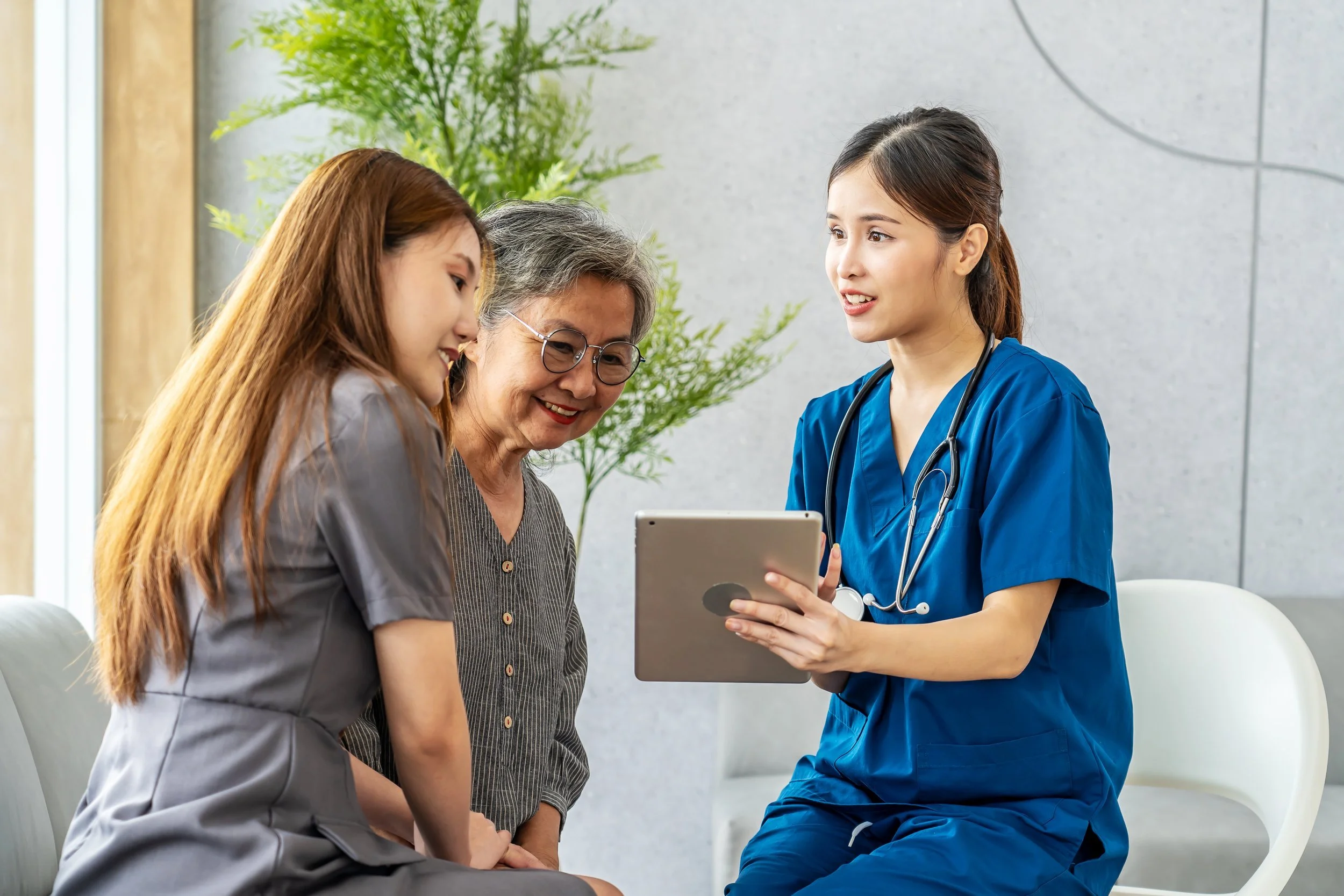 A nurse in blue scrubs shows a tablet to an older woman and a younger woman sitting beside her