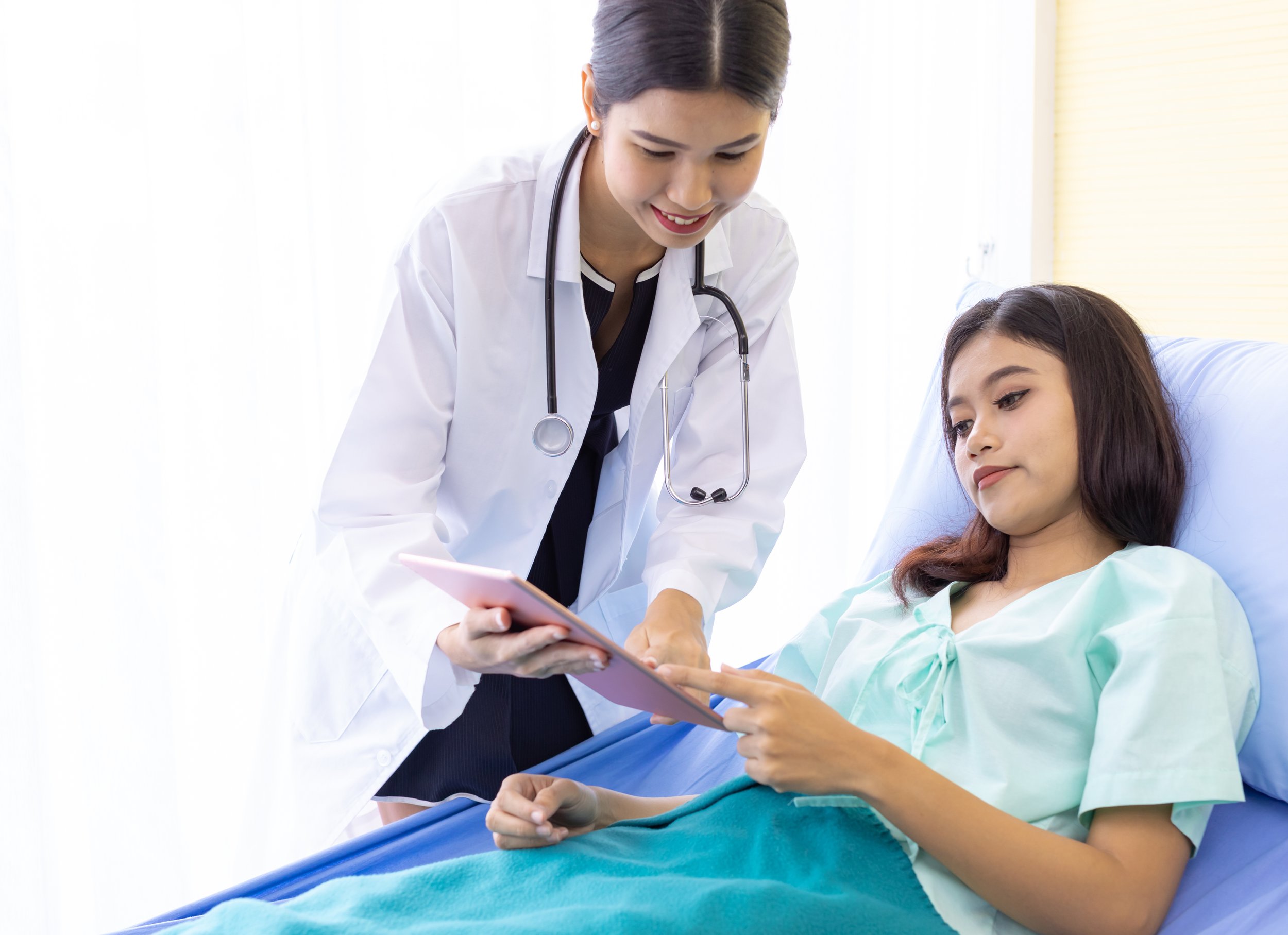 A doctor leans in beside a hospital bed, sharing information on a tablet as the patient reads along.