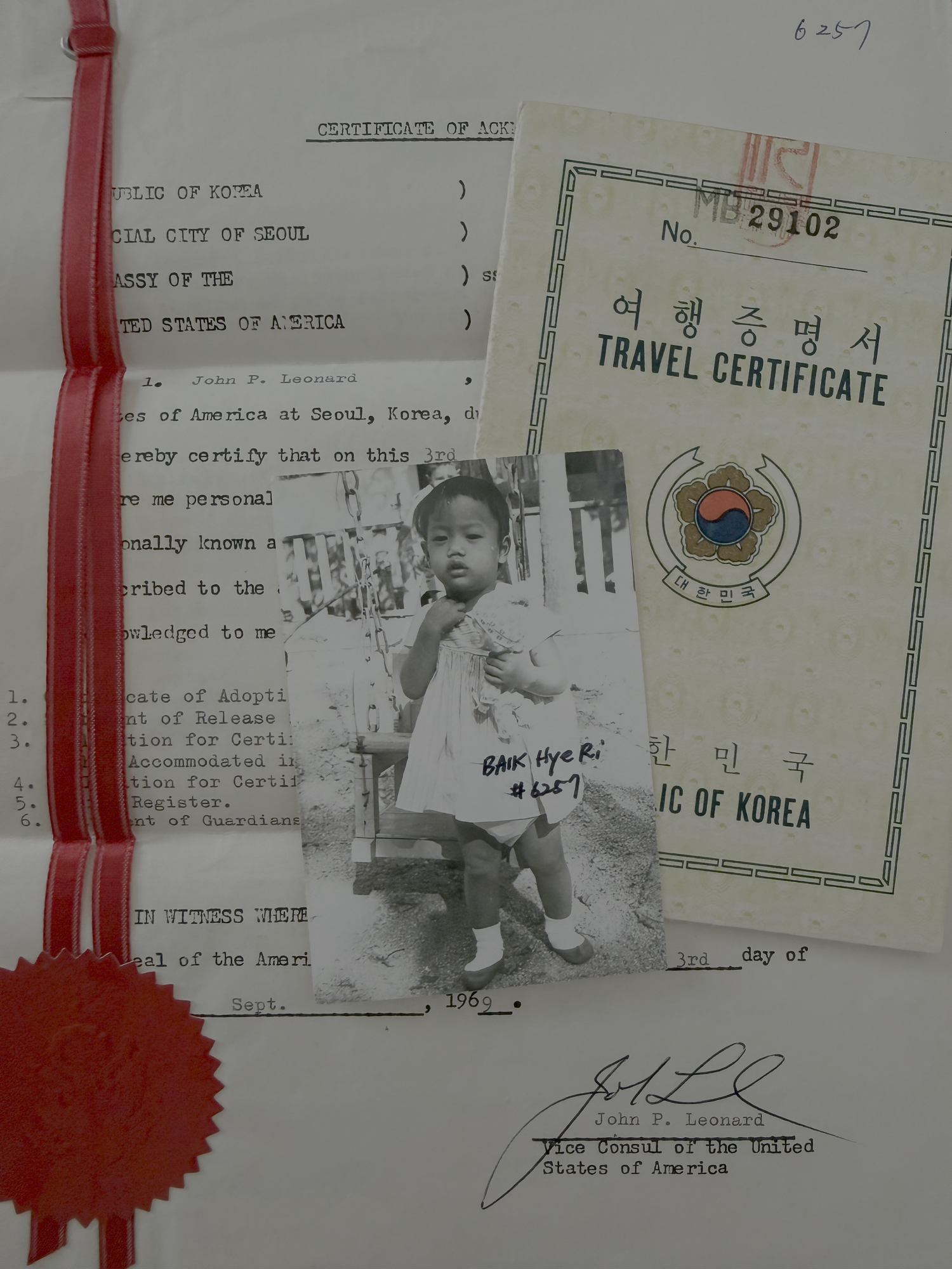 Mia's adoption papers from 1969: a certificate from the U.S. Embassy in Seoul, her Korean travel certificate, and a photo of her as a toddler with her name, Baik Hye Ri, written on it.