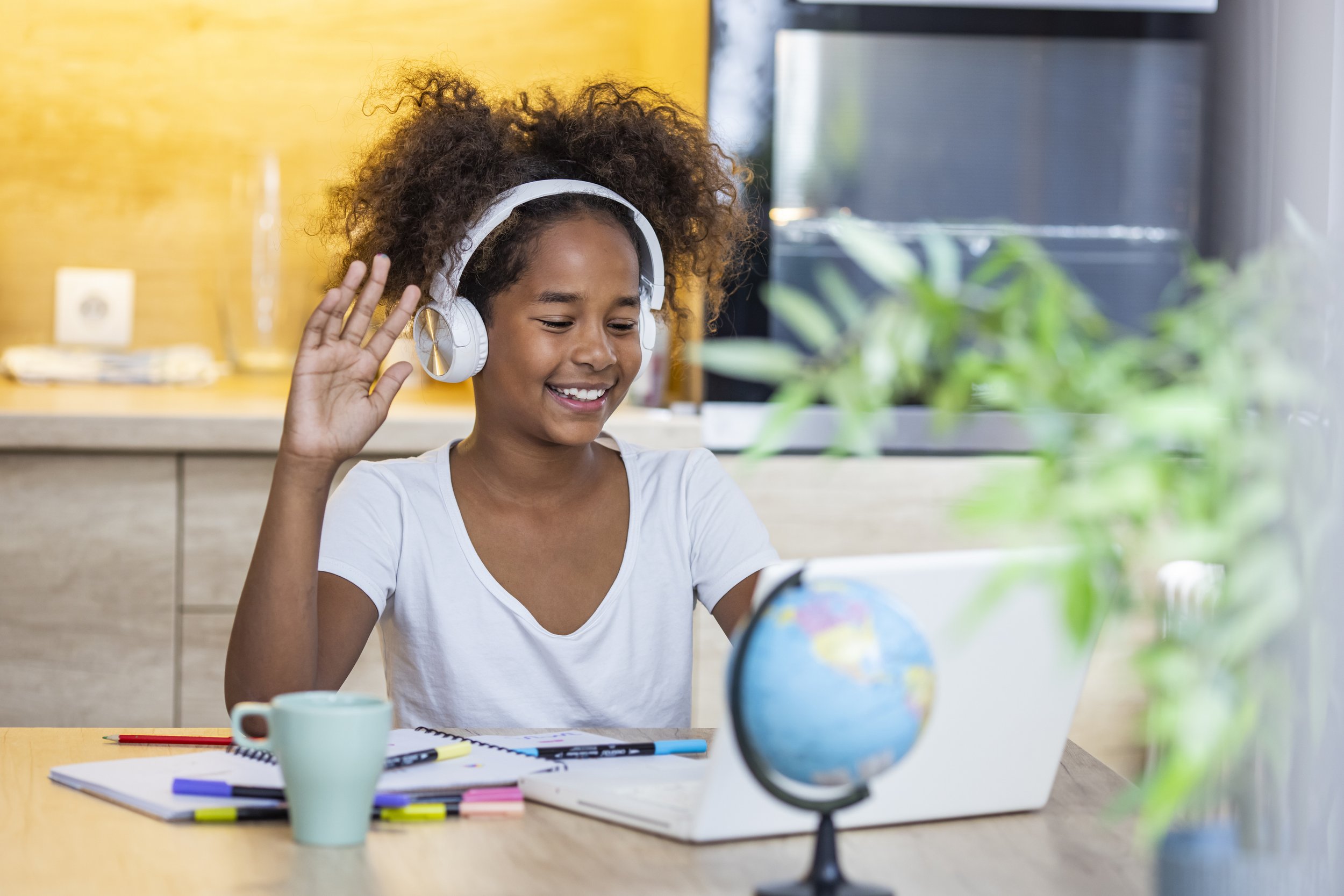 A young woman wearing headphones smiles and waves at her laptop during a video class, with a small globe on the desk beside her.