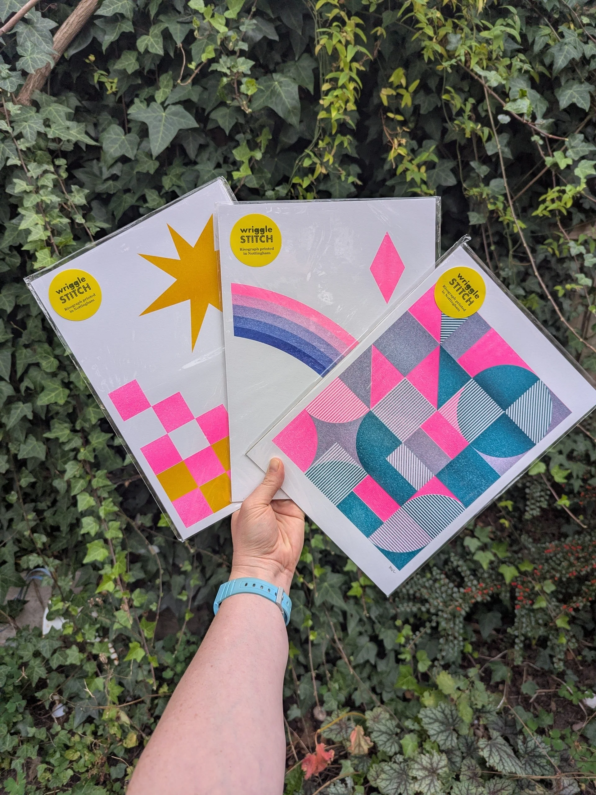 A selection of colourful, abstract prints featuring geometric shapes such as squares, triangles, circles and arcs. The prints are in vibrant colours, pink, yellow, blue, purple and teal. They are packaged ready for sale and held by a hand.