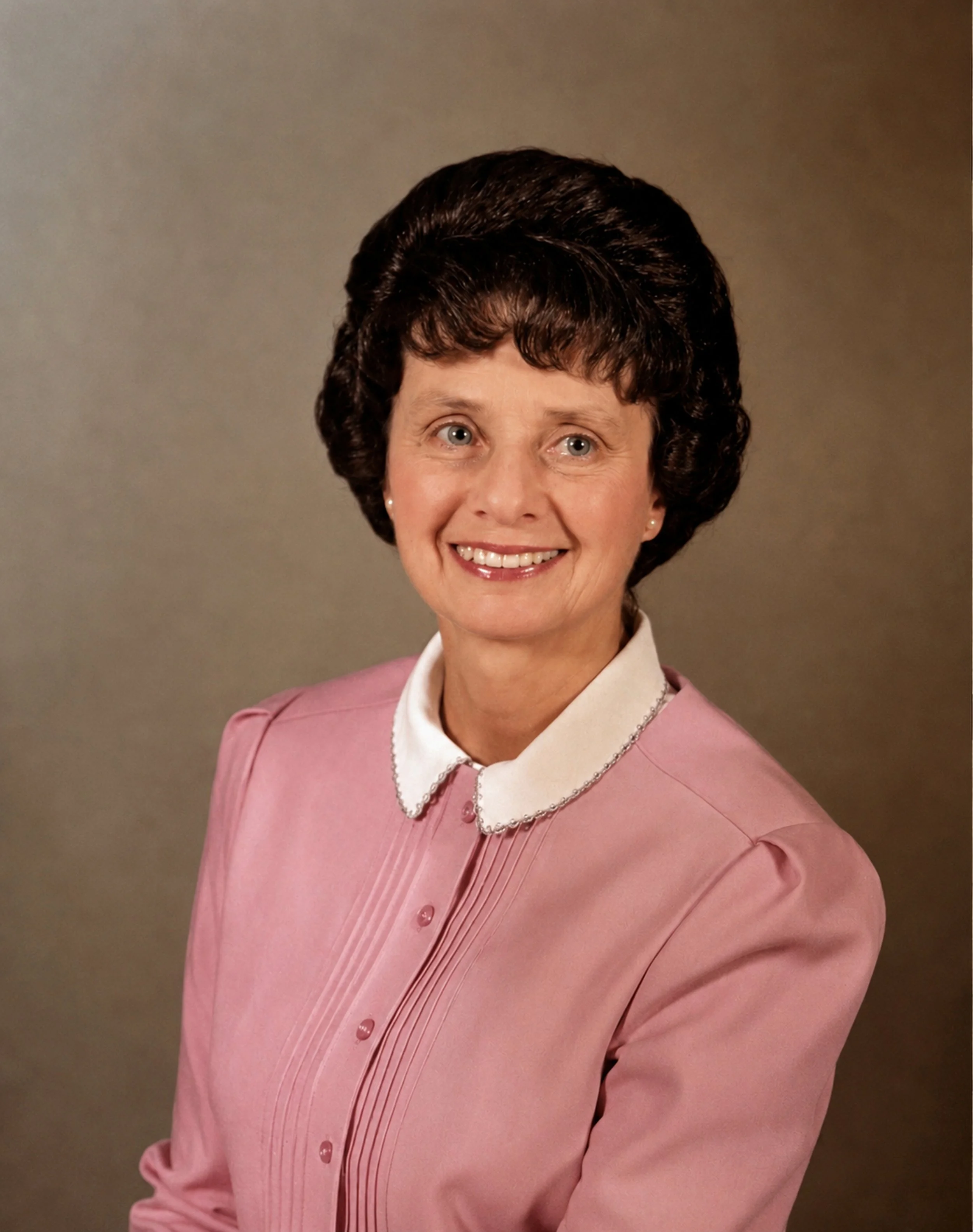 A woman in a pink blazer with a white collar, smiling, against a neutral background.