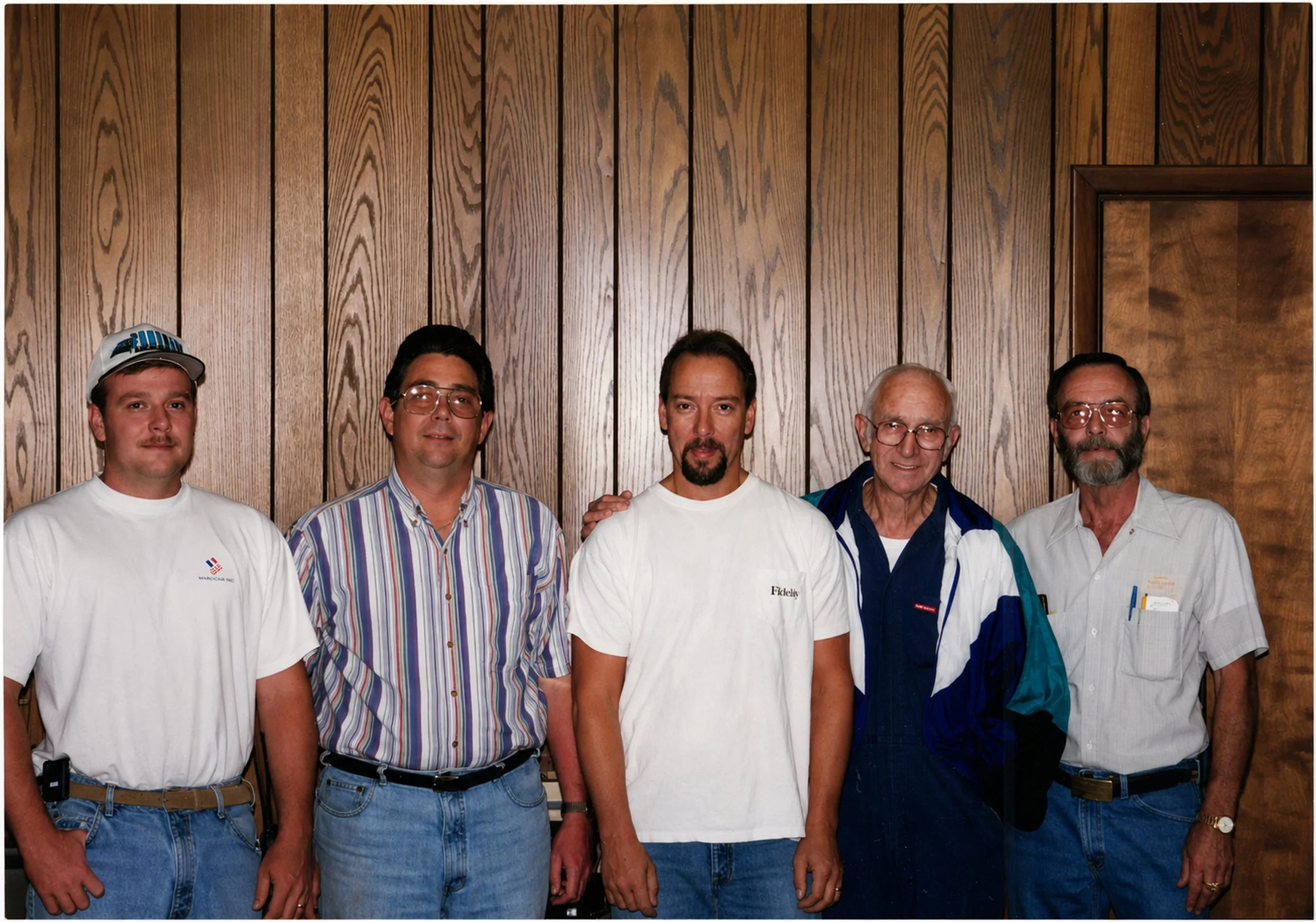 Six men standing together against a wooden paneled wall, dressed casually, with some wearing glasses and sporting various hairstyles and facial hair.