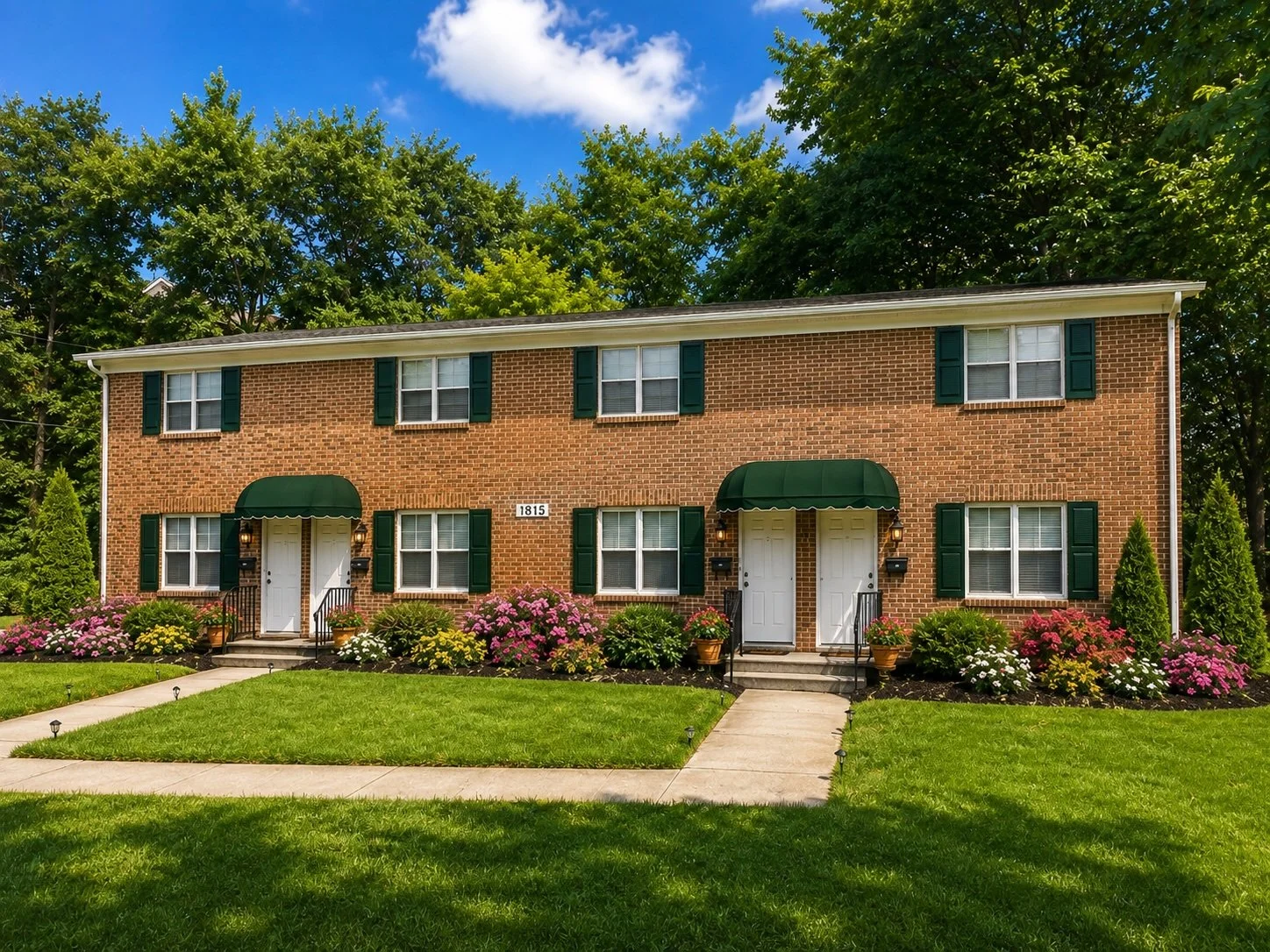 Two identical brick townhouses with green shutters and awnings, white front doors, on a well-manicured lawn with flower beds and bushes, under a blue sky.