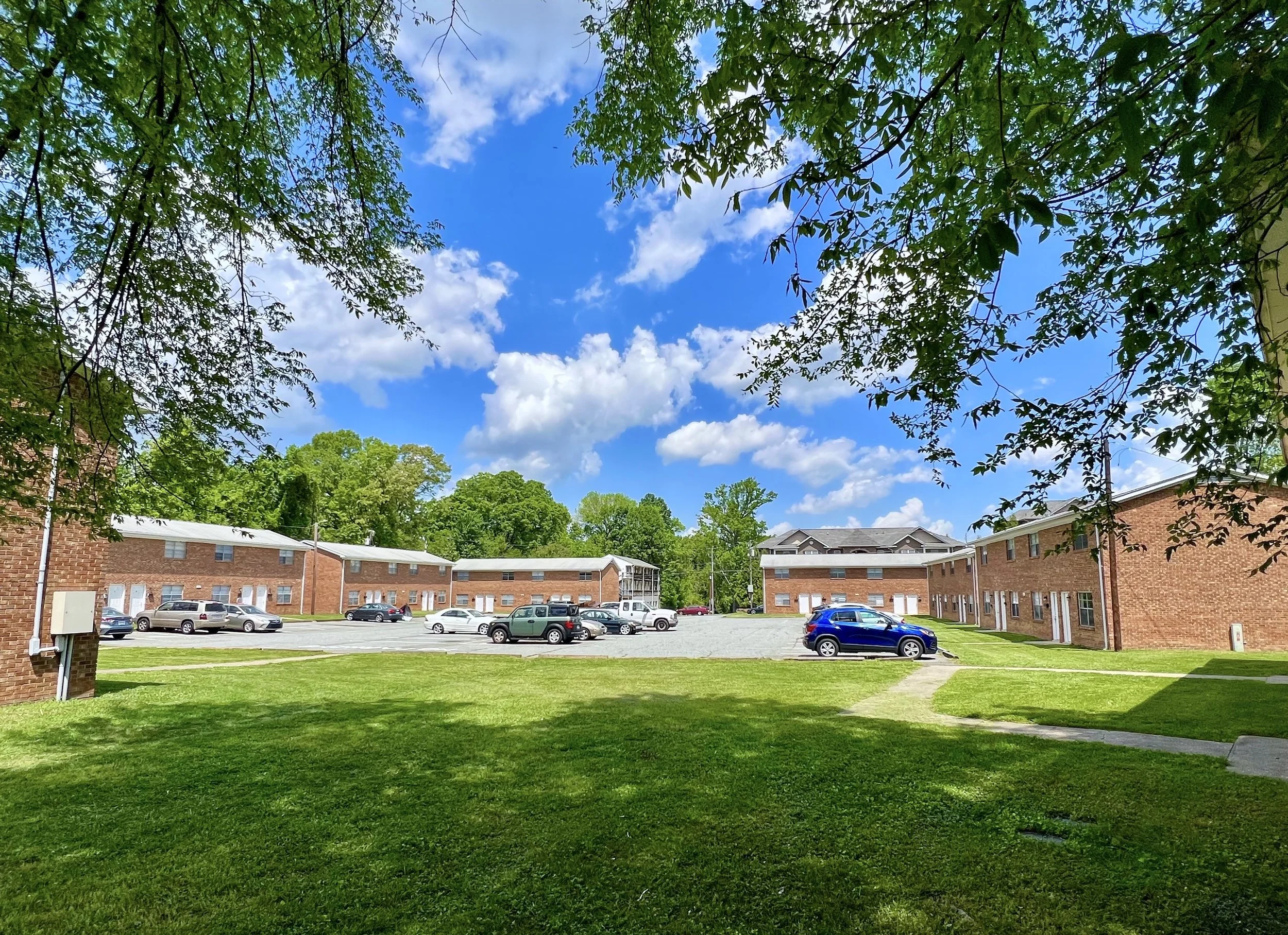 An apartment complex with red brick buildings, a parking lot with several cars, and a grassy area with trees. The sky is partly cloudy with blue sky and white clouds.