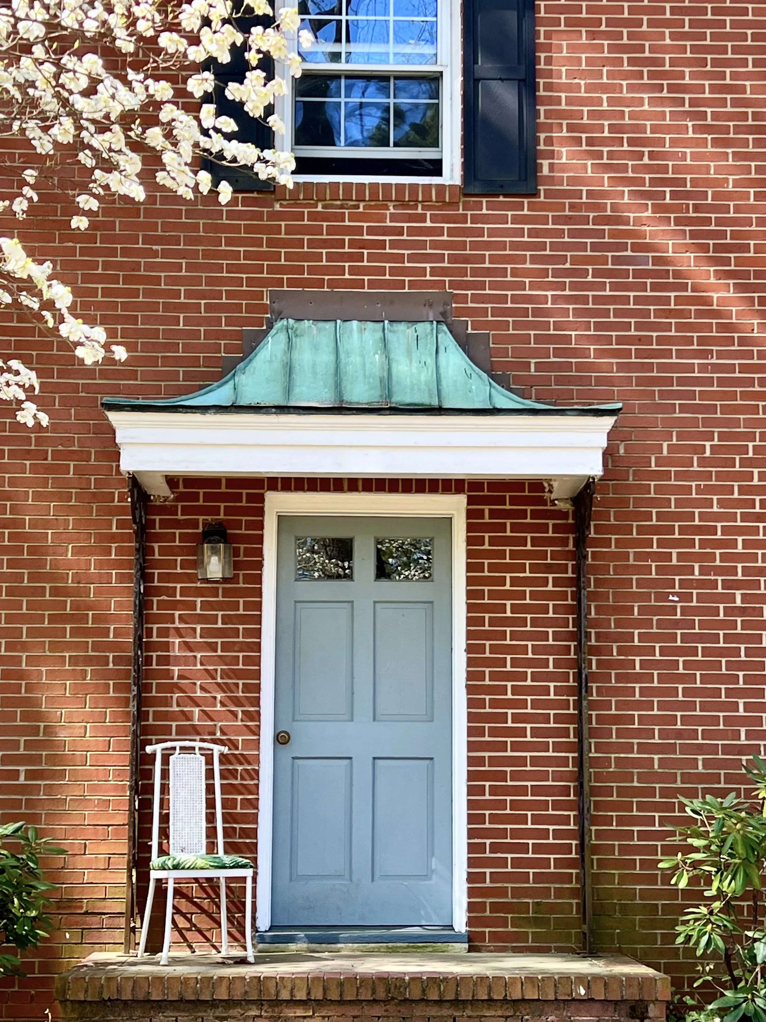 A brick house front with a light blue door, a small porch with a brick step, a white chair with a green cushion, and a blooming tree with white flowers to the left.