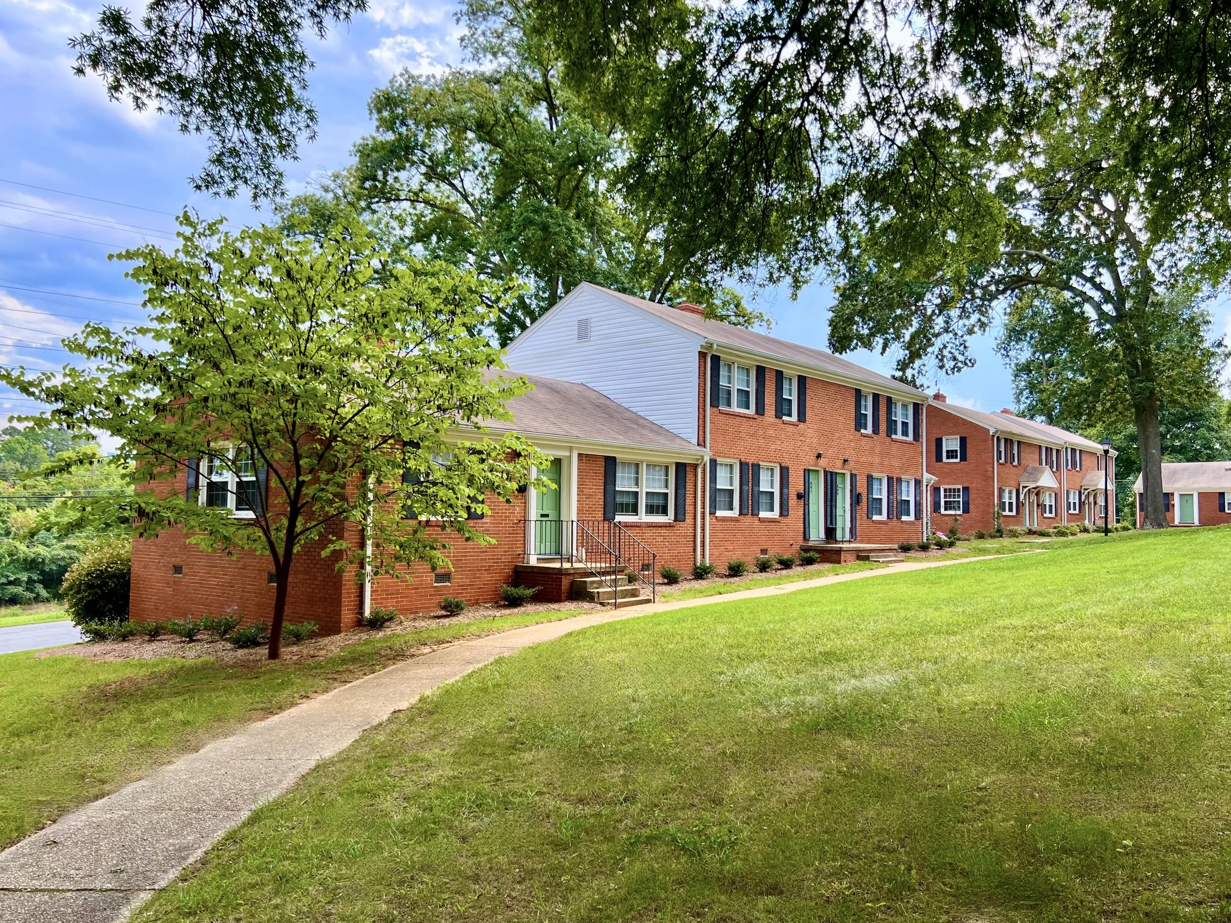 Row of brick townhouses with colorful doors and steps, surrounded by lush green trees and grass in a suburban neighborhood.