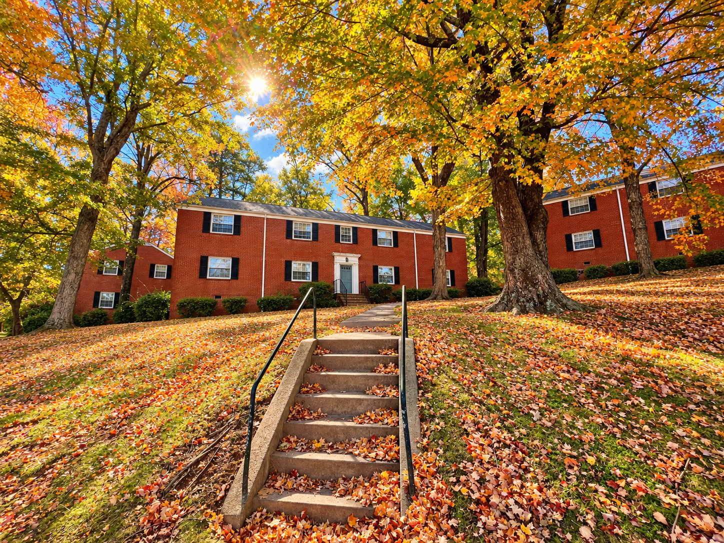 A red brick apartment building surrounded by tall trees with green and yellow autumn leaves. Fallen leaves cover the grassy hill and stairs leading to the entrance. The sky is blue with some clouds, and sunlight filters through the trees.