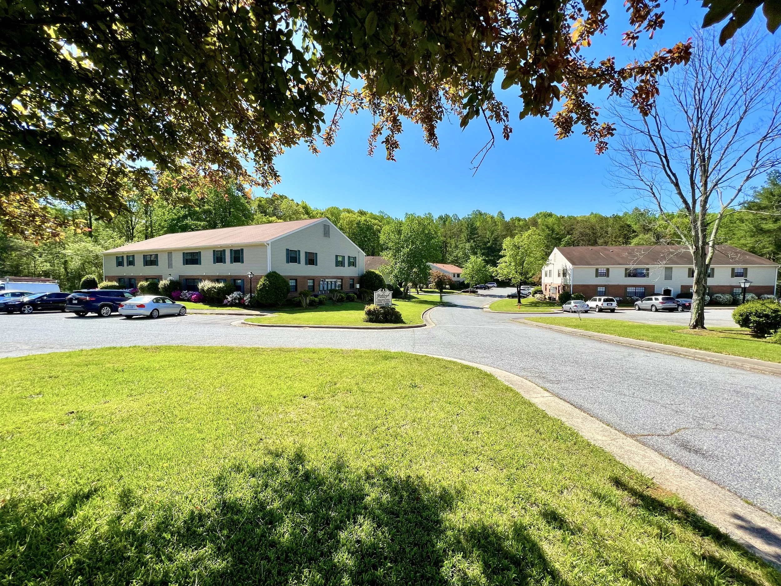 A residential complex with white multi-unit buildings, parking lot, green trees, and a clear blue sky.