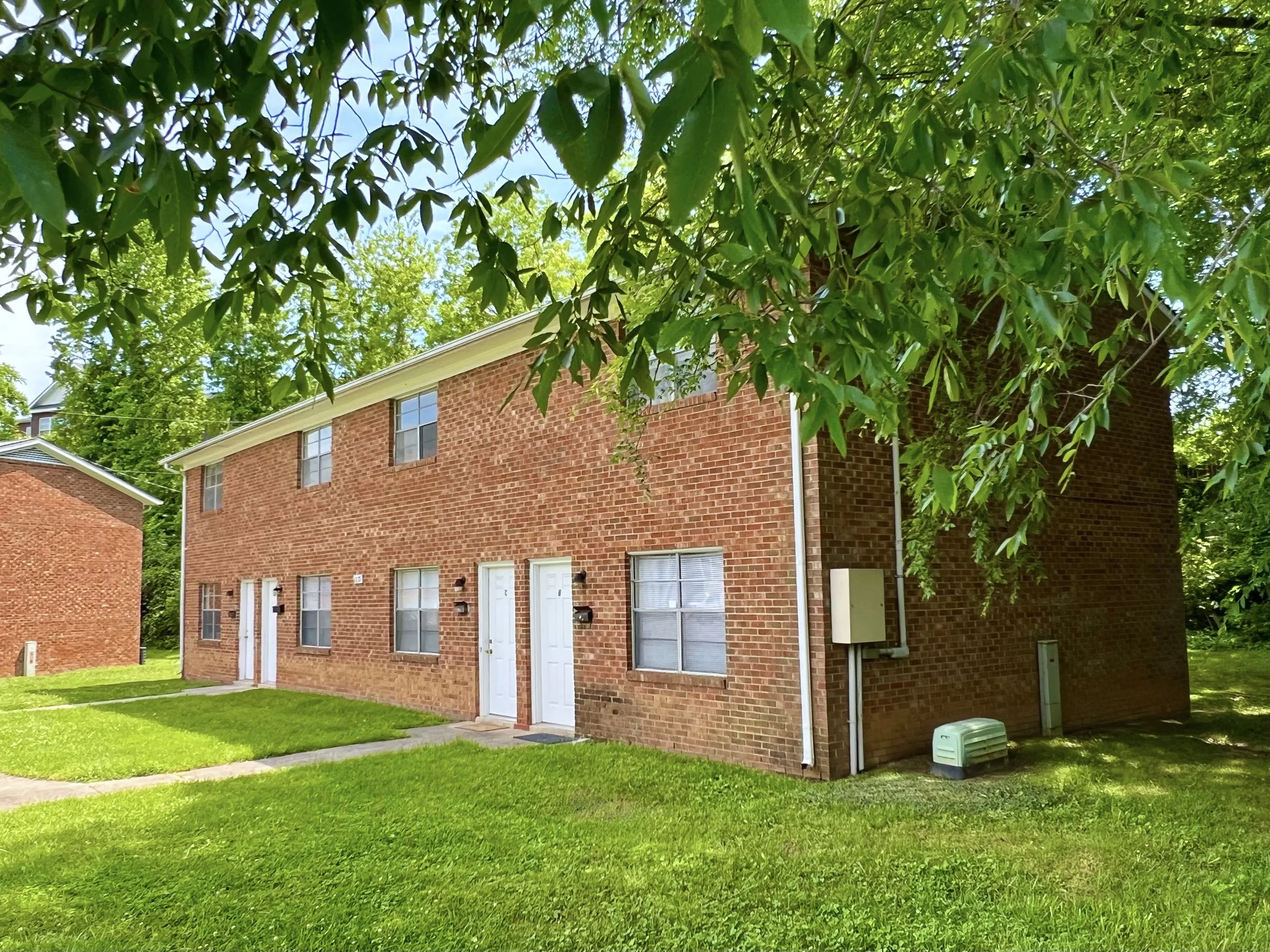 The image shows a row of brick apartment buildings with white doors and windows, surrounded by well-maintained green lawns. There are large leafy trees in the foreground, partially obscuring the buildings and providing shade. The sky is partly cloudy with patches of blue.