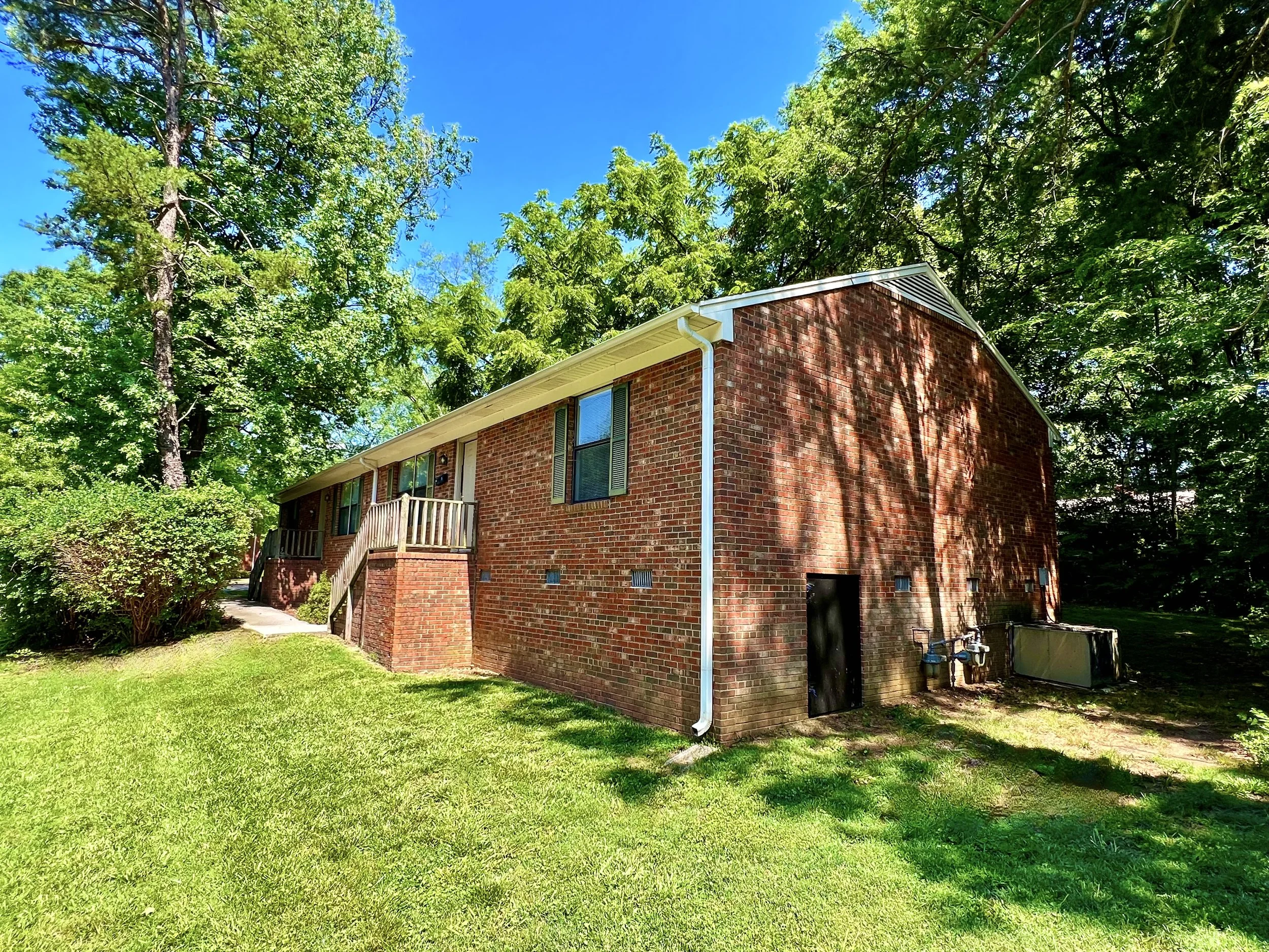 A two-story red brick house with several windows, a small wooden staircase leading to a door, surrounded by lush green trees and grass under a bright blue sky.