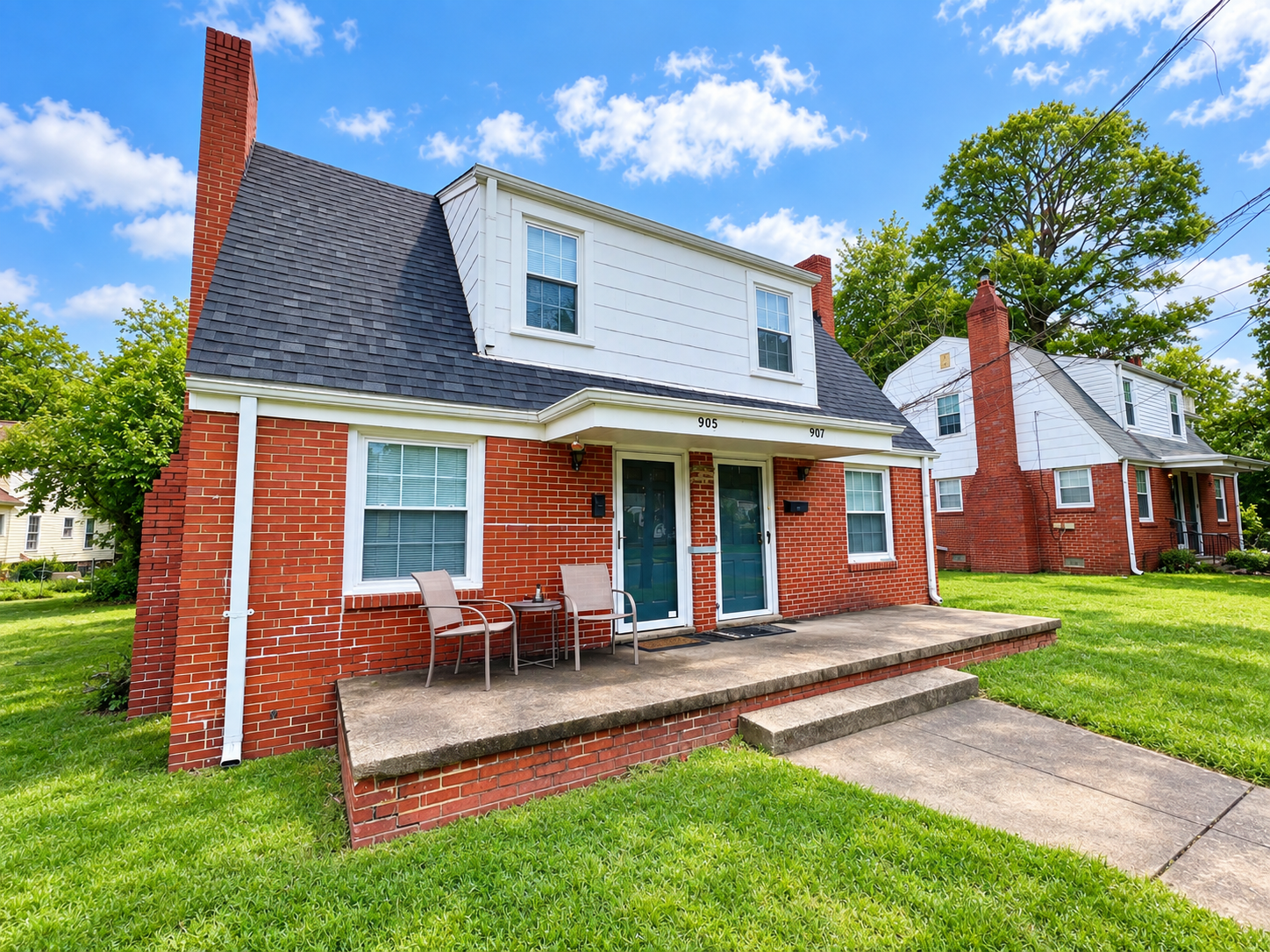 A brick suburban house with a small concrete patio, two chairs, a small table, and a door, under a blue sky with some clouds.
