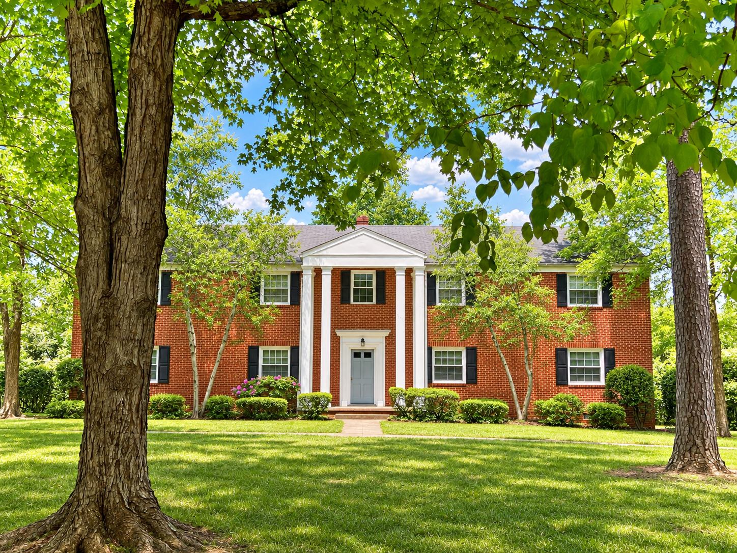 A large brick house with black shutters, centered pillars, and a gray front door, surrounded by green trees and well-maintained lawn, under a partly cloudy sky.
