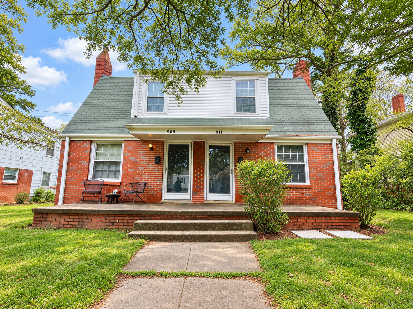 A two-story brick house with a green shingled roof, front porch, and steps leading up to it, surrounded by green grass and trees, under a partly cloudy blue sky.