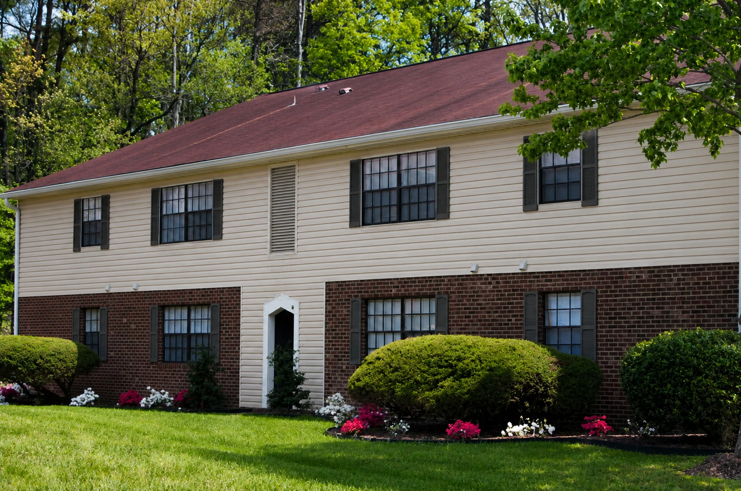 A two-story house with beige vinyl siding, black window shutters, a red shingle roof, and a white front door. The house has a brick foundation on the lower level, surrounded by a well-kept lawn with bushes and colorful flowers.