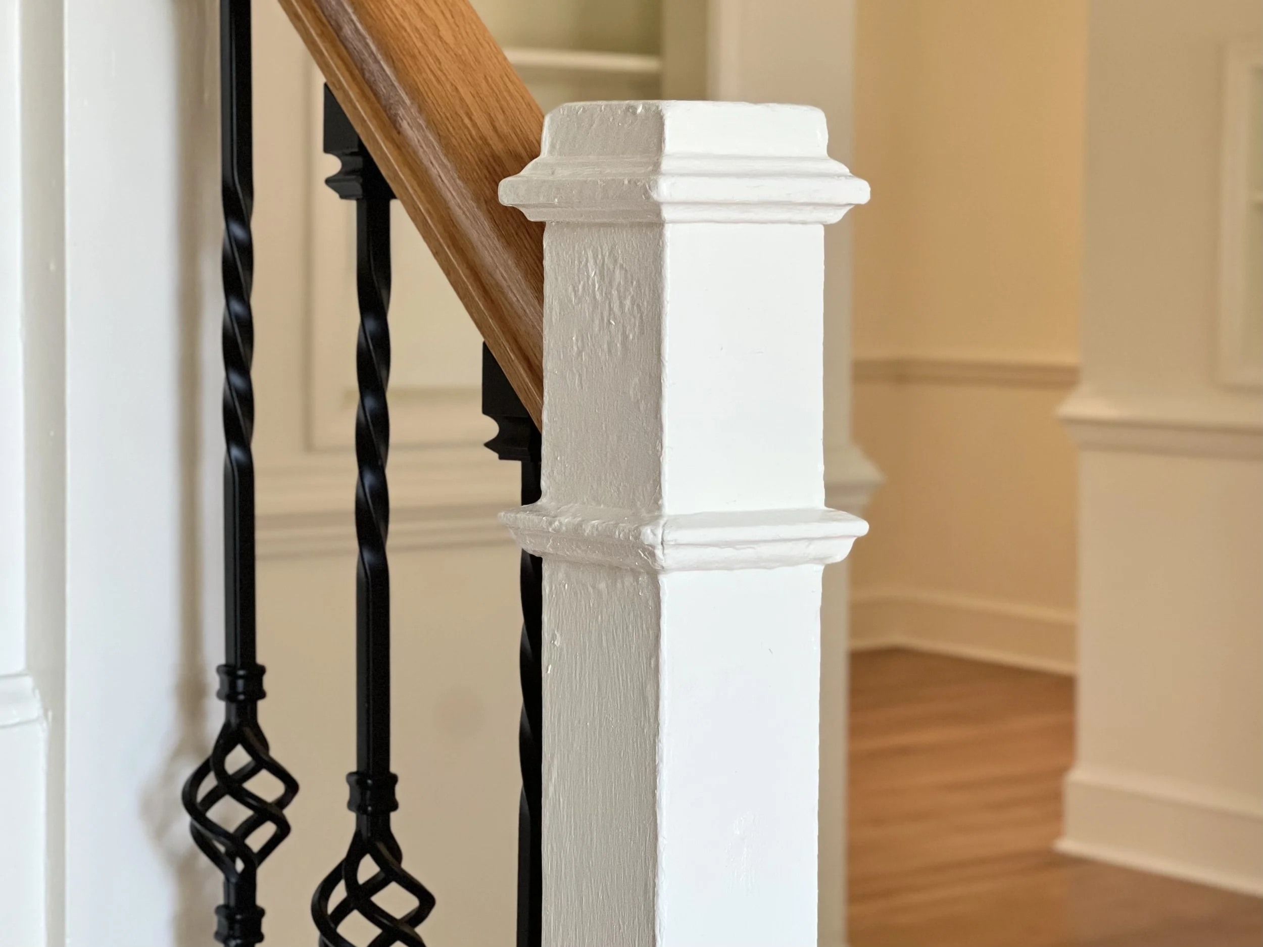 Close-up view of a white painted stair railing post with decorative trim, wood handrails, and black twisted metal balusters in a house interior.