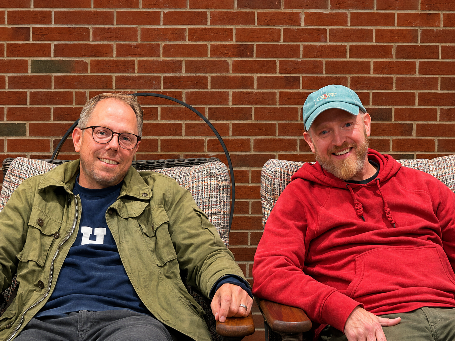 Two men sitting on chairs against a brick wall, smiling at the camera. The man on the left has glasses, a beard, and is wearing a green jacket over a navy shirt. The man on the right has a beard and is wearing a blue cap with the word 'CREW' and a red hoodie.