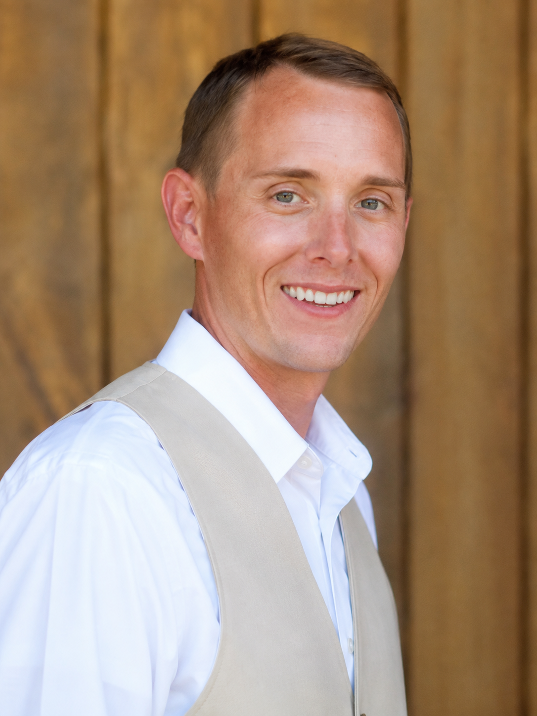 Smiling man in white dress shirt with beige vest in front of wooden background.