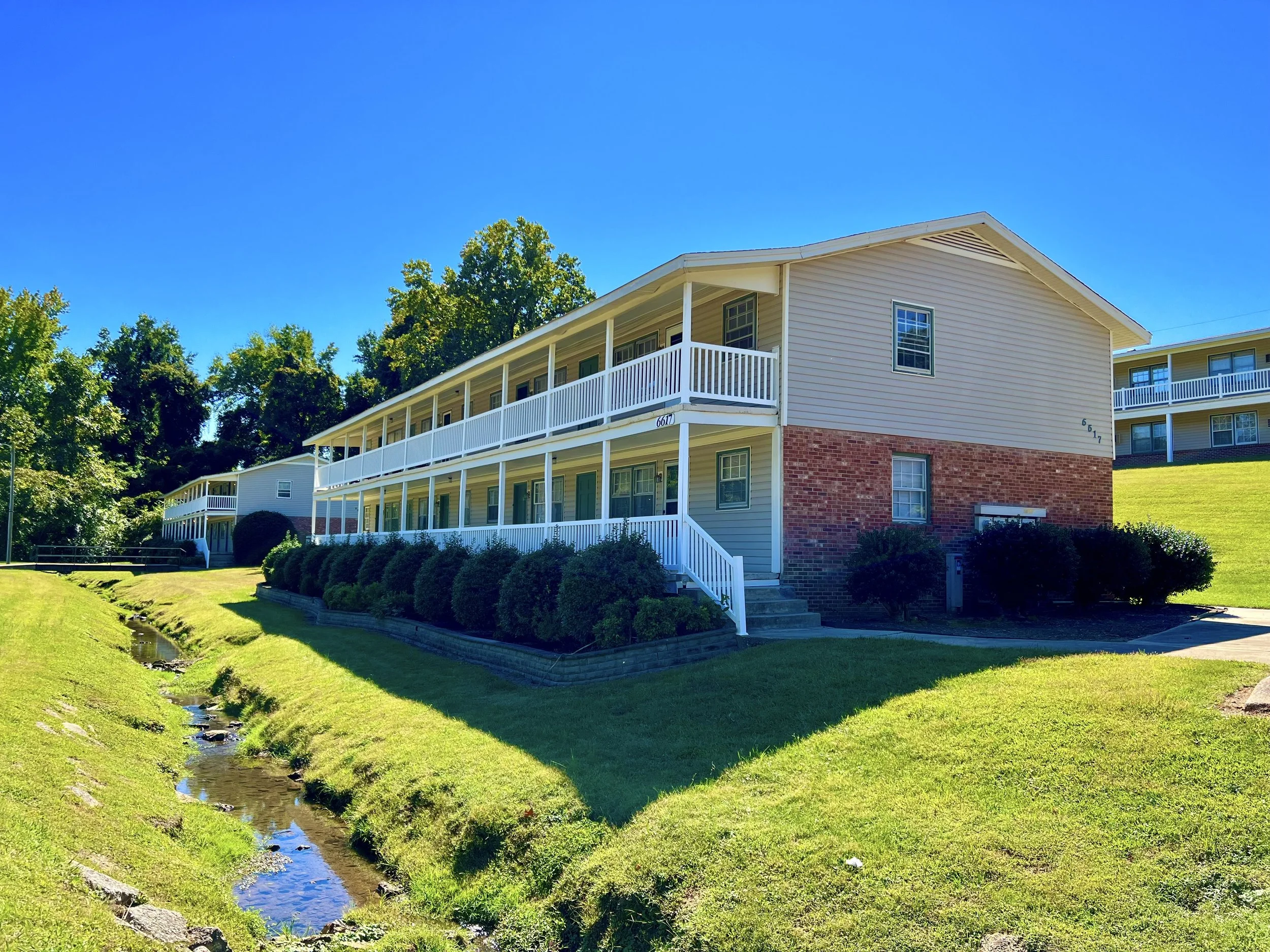 Multi-story residential buildings with white railings and beige siding, surrounded by green grass, trees, and a small creek, under a clear blue sky.