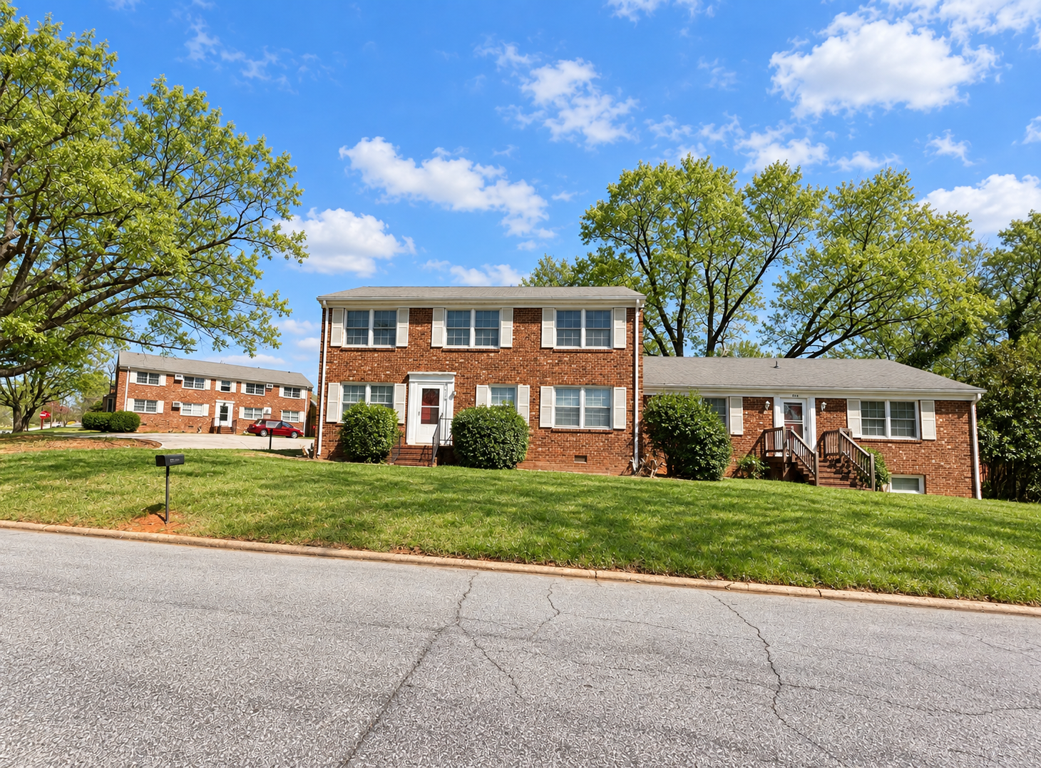A two-story brick house with white window shutters and a small front porch, surrounded by a green lawn and trees, under a blue sky with scattered clouds.
