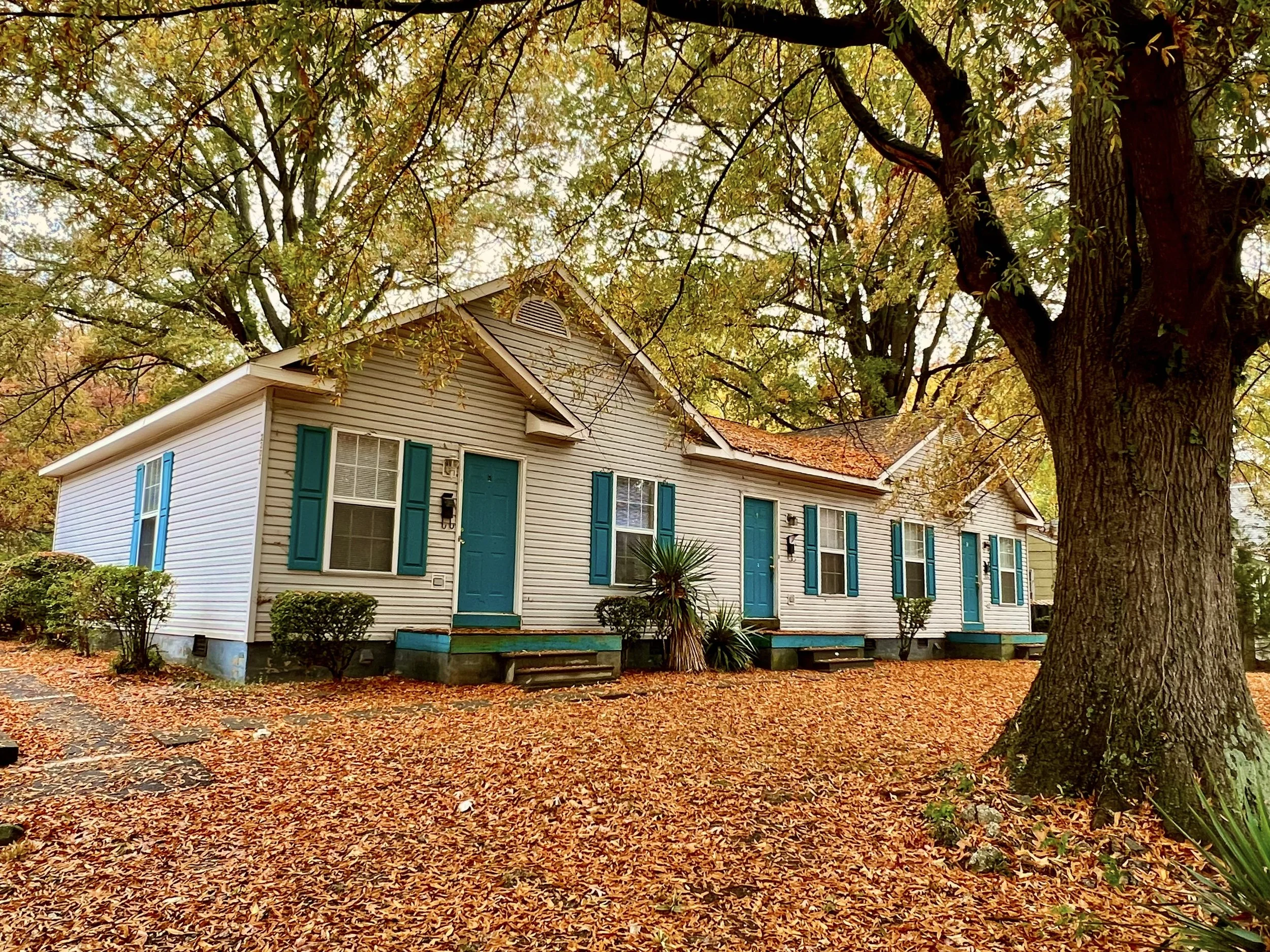 A two-story house with white siding, teal shutters, and a teal front door, surrounded by large trees with green and yellow leaves. The ground is covered with fallen leaves, indicating autumn season.